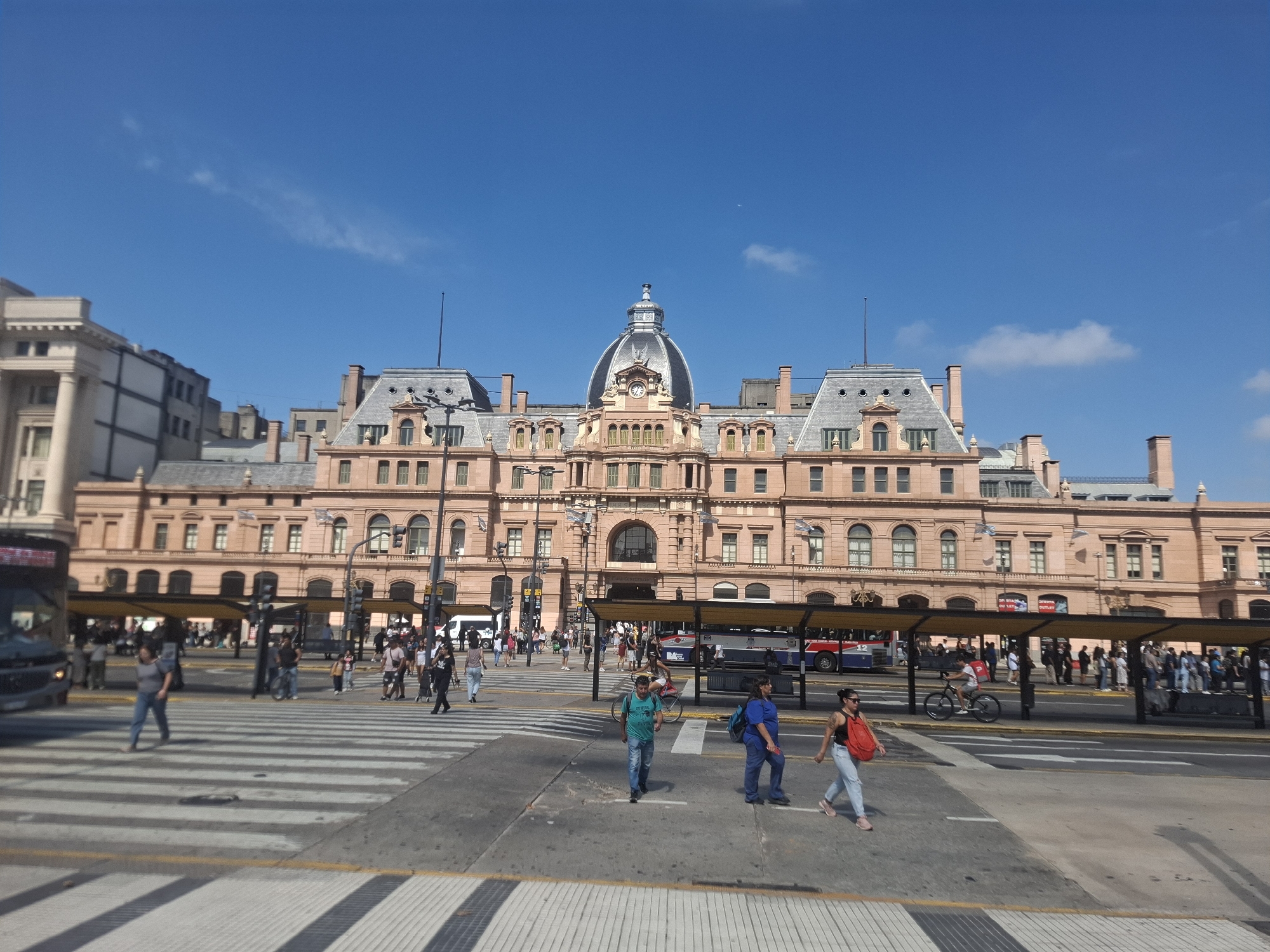 The image depicts a daytime street scene in Buenos Aires, Argentina, focused on the Retiro railway station. The station, a large, pinkish building with a prominent dome and intricate architectural details, dominates the background. In the foreground, a busy intersection with crosswalks is visible, with pedestrians crossing the street. Some are walking alone, while others are in small groups. A cyclist is riding across the street. Yellow bus shelters are located alongside the road, with people waiting inside. The sky is clear and blue. The overall impression is of a bustling urban environment.