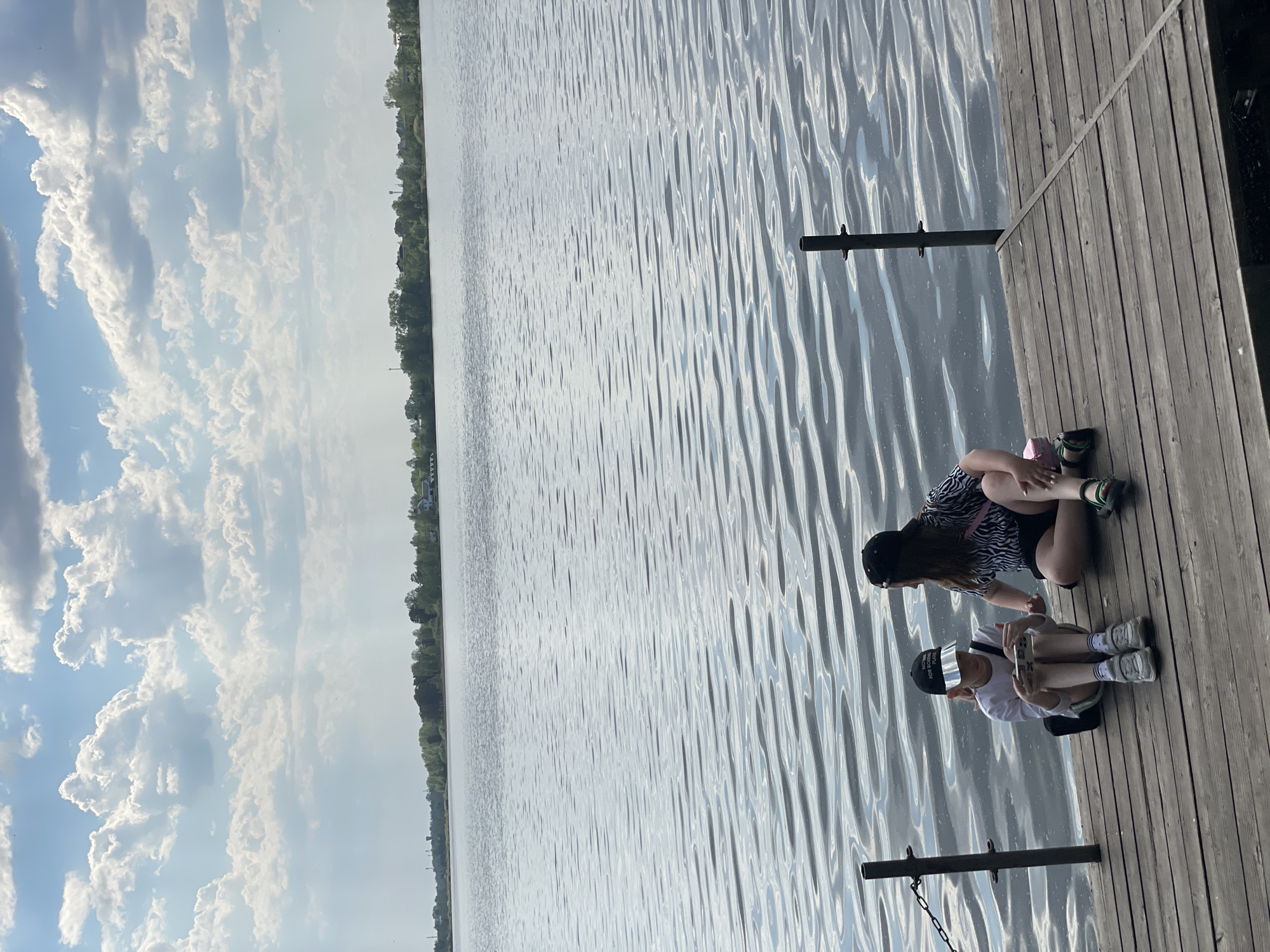 Two individuals are seated on a wooden dock extending into a body of water. The person closest to the viewer is wearing a white shirt, black shorts, a black baseball cap, and light-colored sneakers, and is holding a cell phone. The other person wears a zebra-striped top, black shorts, and sneakers. The water surface shows ripple patterns. On the horizon opposite the dock, there is a tree line. The sky overhead is partly cloudy. The location is assumed to be Zheleznogorsk, Russia, although this cannot be verified from the image.