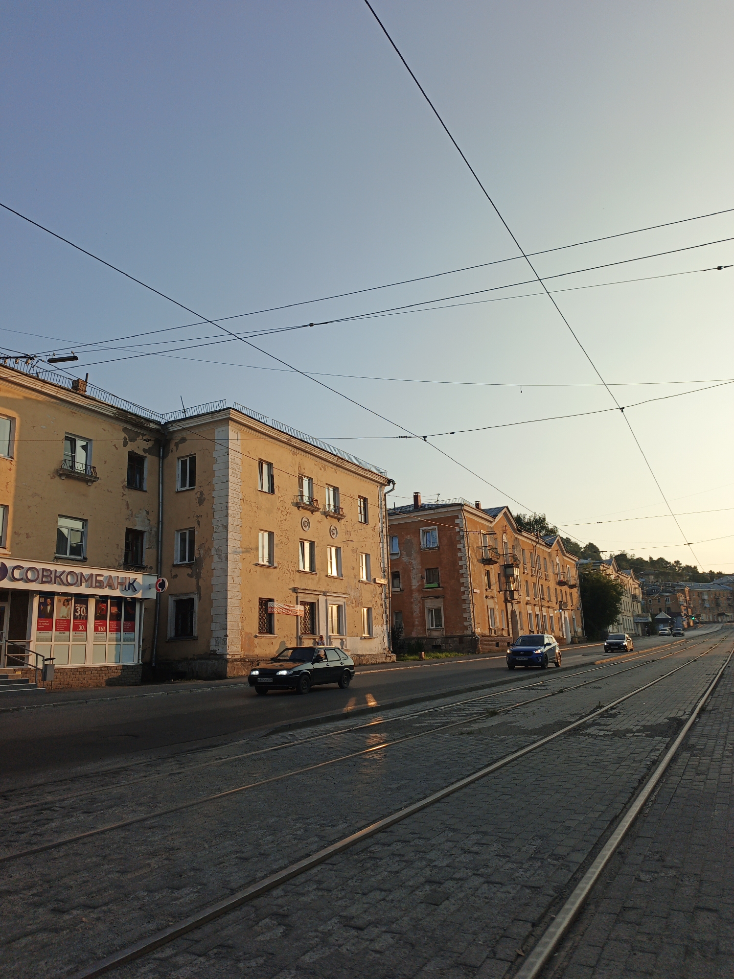 The image captures a street scene in Zlatoust, Russia, during what appears to be late afternoon or early evening, judging by the warm, low-angled sunlight. The composition is dominated by a street with cobblestone paving and tram tracks running parallel to the buildings. On the left side of the street, a pale yellow, multi-story building features a storefront with a sign that reads "СОВКОМБАНК" (Sovcombank). Several cars are visible on the road, including a dark sedan in the foreground and other vehicles further down the street. The buildings lining the street are older, with stucco facades in shades of yellow and orange, some showing signs of wear. Overhead, numerous electrical wires crisscross the sky, likely for trams or other urban infrastructure. The sky is clear and light blue, transitioning to a warmer hue near the horizon. In the background, a gentle hill rises, with more buildings and trees visible. A few pedestrians can be seen near the entrance of a building on the right side of the street. The overall impression is one of an ordinary urban setting bathed in soft, golden light.