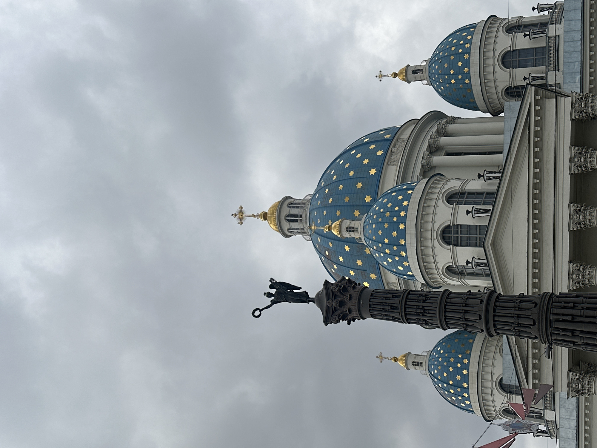 The image depicts a section of the Trinity Cathedral in Saint Petersburg, Russia. The foreground features a dark-colored column topped with a statue of a winged figure holding a wreath. The cathedral's distinctive blue domes adorned with gold stars are visible in the background. The building is white with architectural details like pillars and arched windows. The sky is overcast.