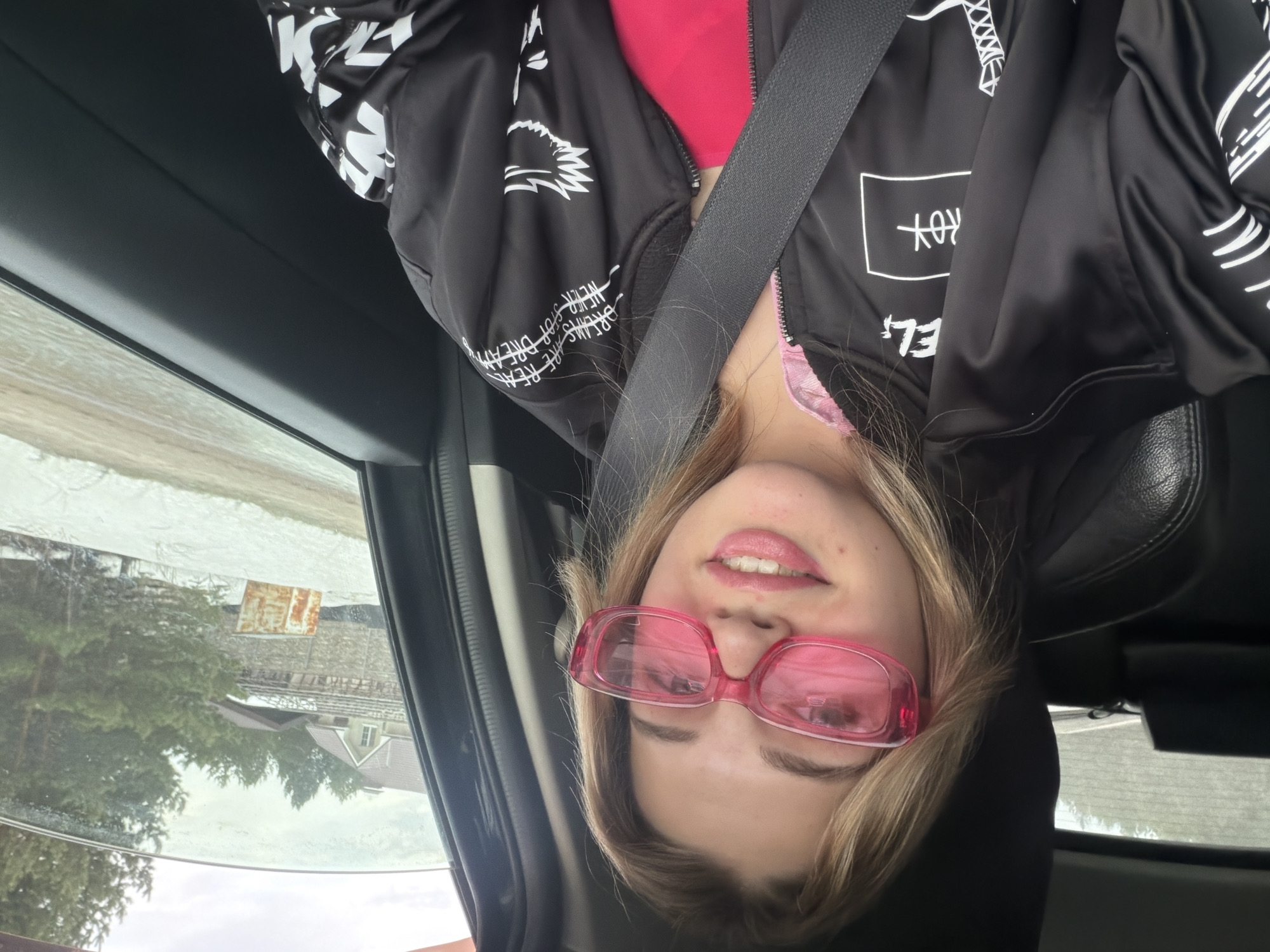 A woman wearing pink sunglasses, a black bomber jacket with white designs, and a pink top is in a car, wearing a seatbelt. Outside the car window, trees and buildings are visible. The image is upside down. Without more information, I cannot verify or locate in Naberezhnye Chelny, Russia.