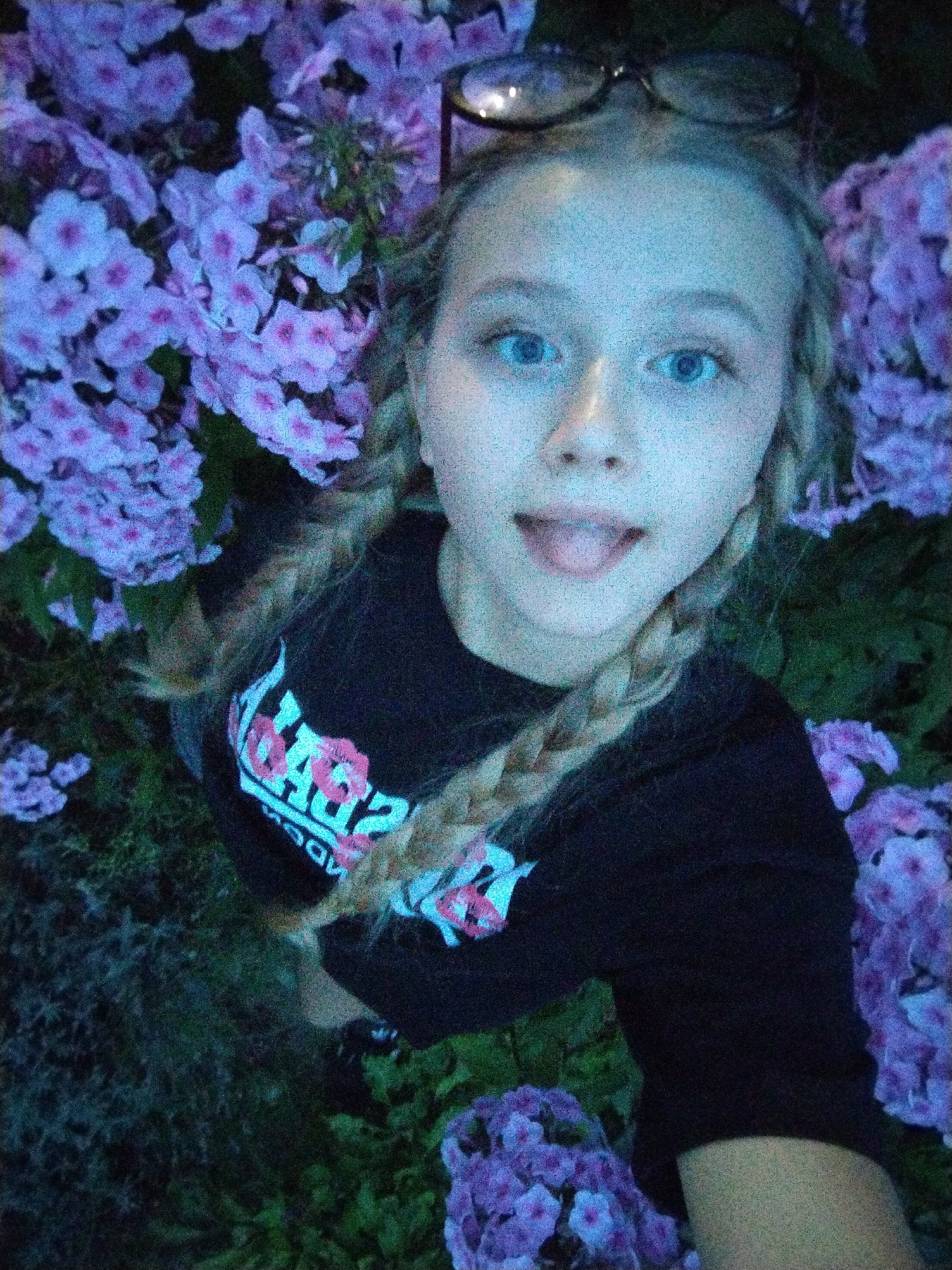 A young person with blonde hair in two braids is pictured in a selfie taken outdoors, surrounded by purple flowers. The person is wearing a black t-shirt with a graphic design that includes the words "SUB..." and "ADD". They have blue eyes and are sticking their tongue out, with glasses perched on their head. The scene is illuminated by artificial light, giving the image a grainy, blueish tint. The setting appears to be a garden or park area with dense foliage. No specific landmarks of Bialynichy, Belarus, are visible in the image.