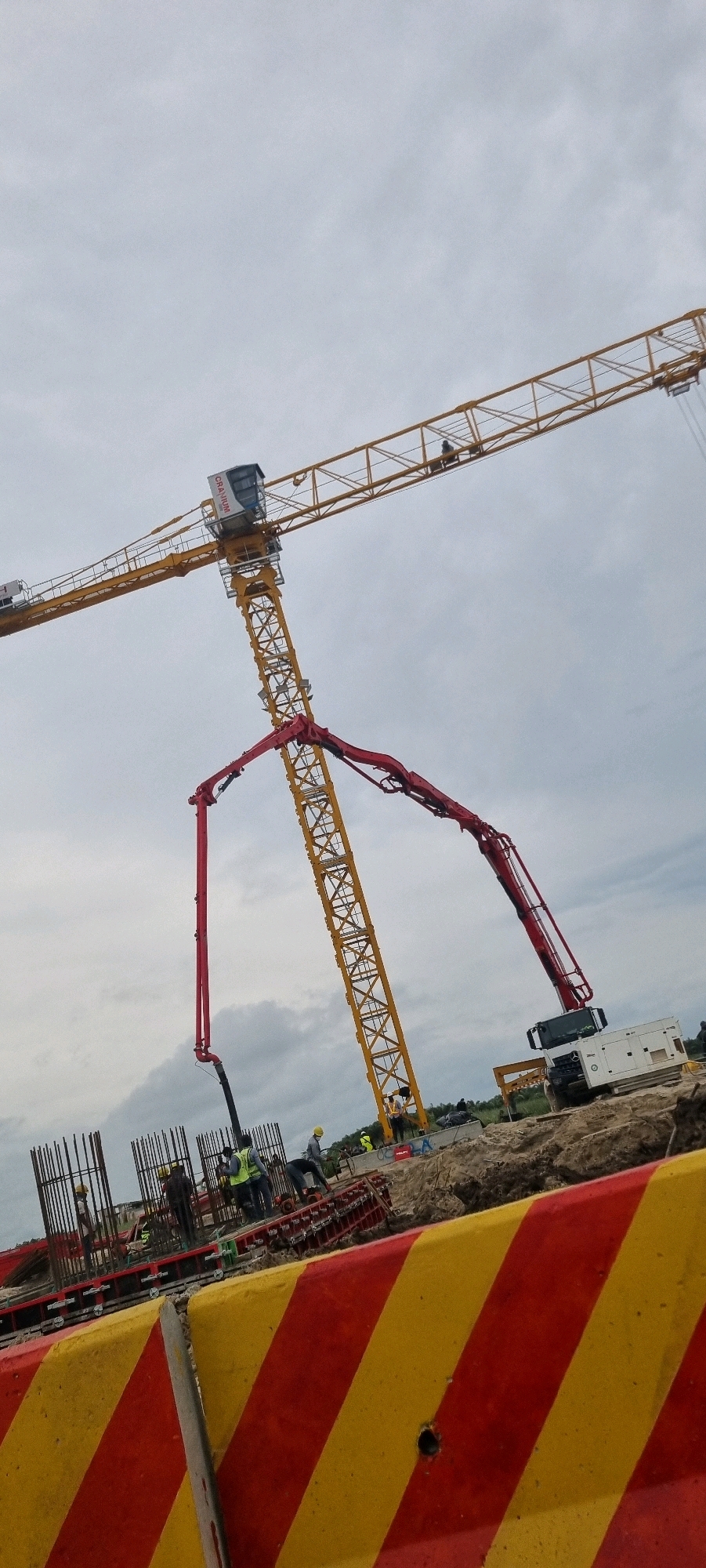 In Mopo Onijebu, Nigeria, a construction site is captured under an overcast sky. A tall yellow tower crane dominates the scene, its boom extended horizontally and then angled downwards. Attached to the crane is a red concrete pump boom, positioned to deliver material. Beneath the crane, a white truck with a generator is parked. Several construction workers, some wearing high-visibility vests and hard hats, are visible near the base of the crane and around rebar structures. In the foreground, a barrier with alternating red and yellow diagonal stripes marks a boundary on the dusty ground. The overall environment suggests active construction work.