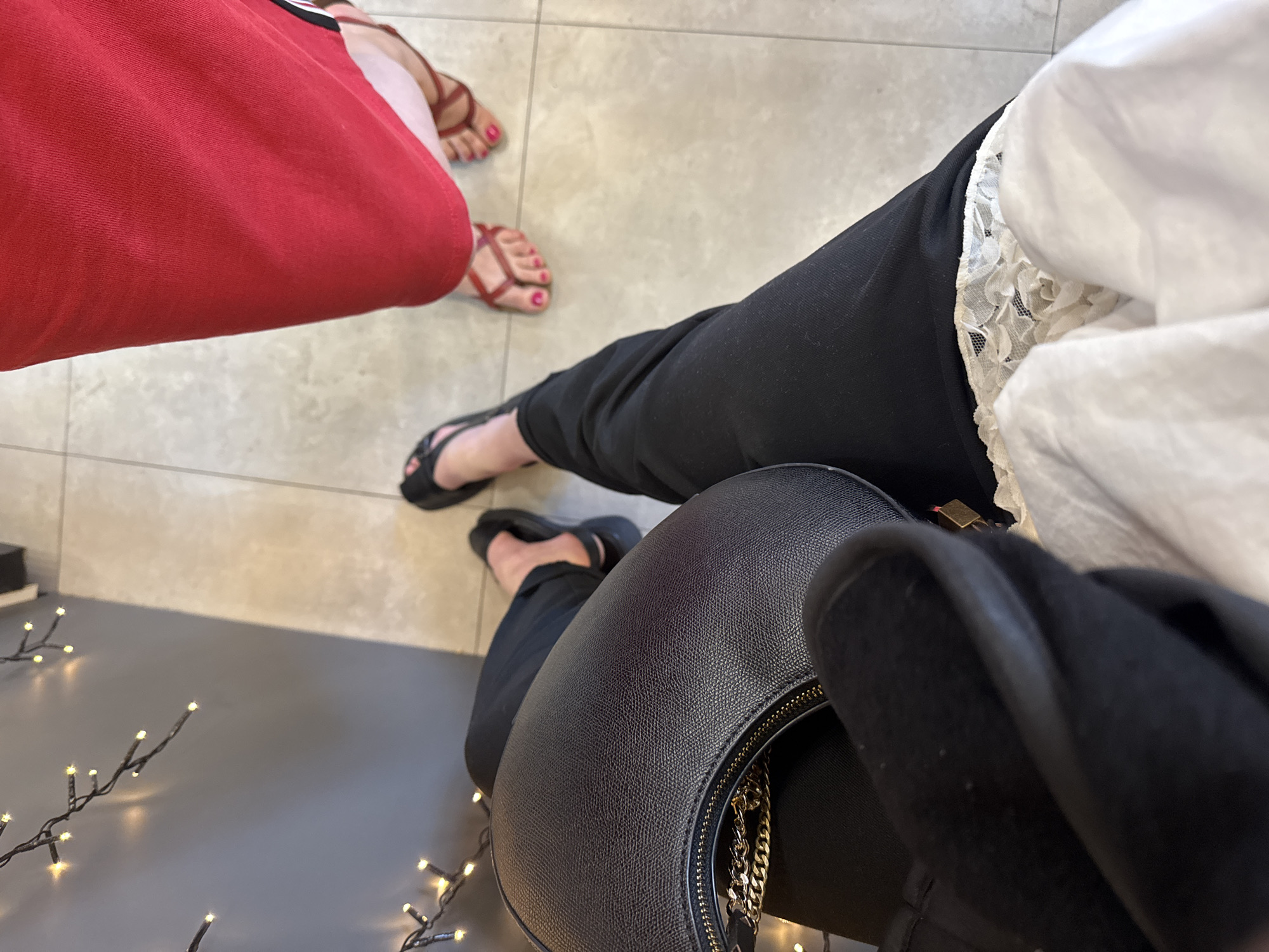 Viewed from above, the image shows two individuals and their surroundings. One individual, partially obscured by a large red bag, wears brown strappy sandals with pink toenail polish. Another person is visible, wearing black trousers, a white top with lace detail, and black flip-flops. A black handbag with a gold chain detail rests in their lap. The scene includes the flooring, which transitions from light-colored square tiles to a dark, possibly polished surface where decorative lights are arranged. The location is in the city of Unknown, Unknown.