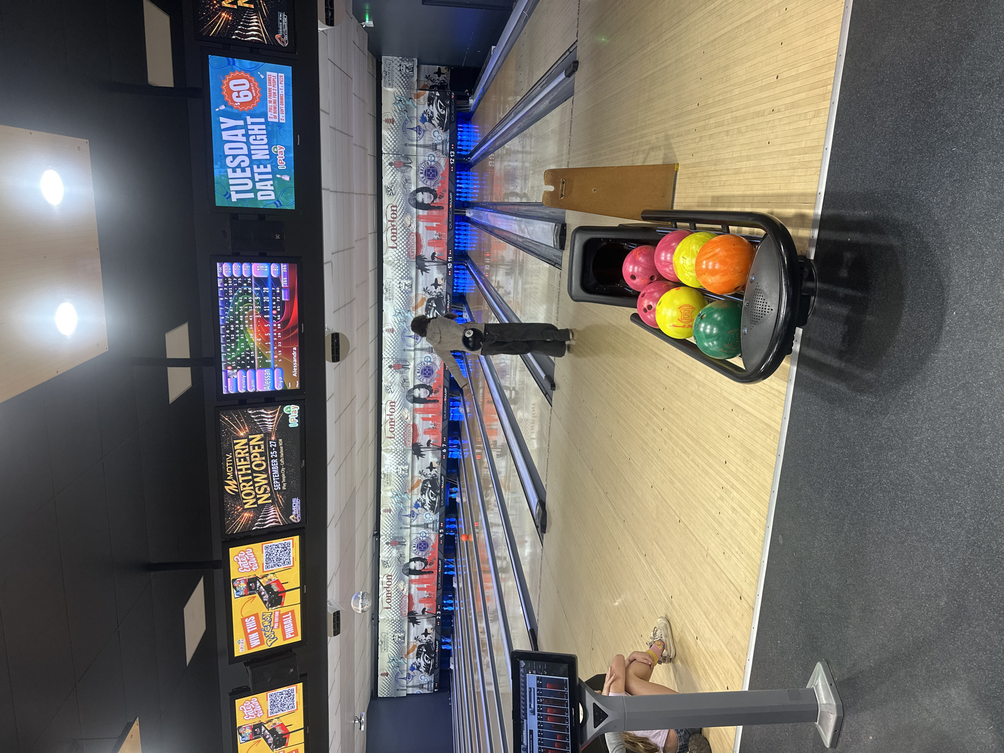 The image shows the interior of a bowling alley. A young person is in the middle of the lane, about to bowl. A ball return rack is in the foreground, filled with several bowling balls of various colors. Large screens above the lanes display advertisements and scores. The bowling alley is decorated with imagery of London and New York. The bowling alley is located in Coffs Harbour, Australia.