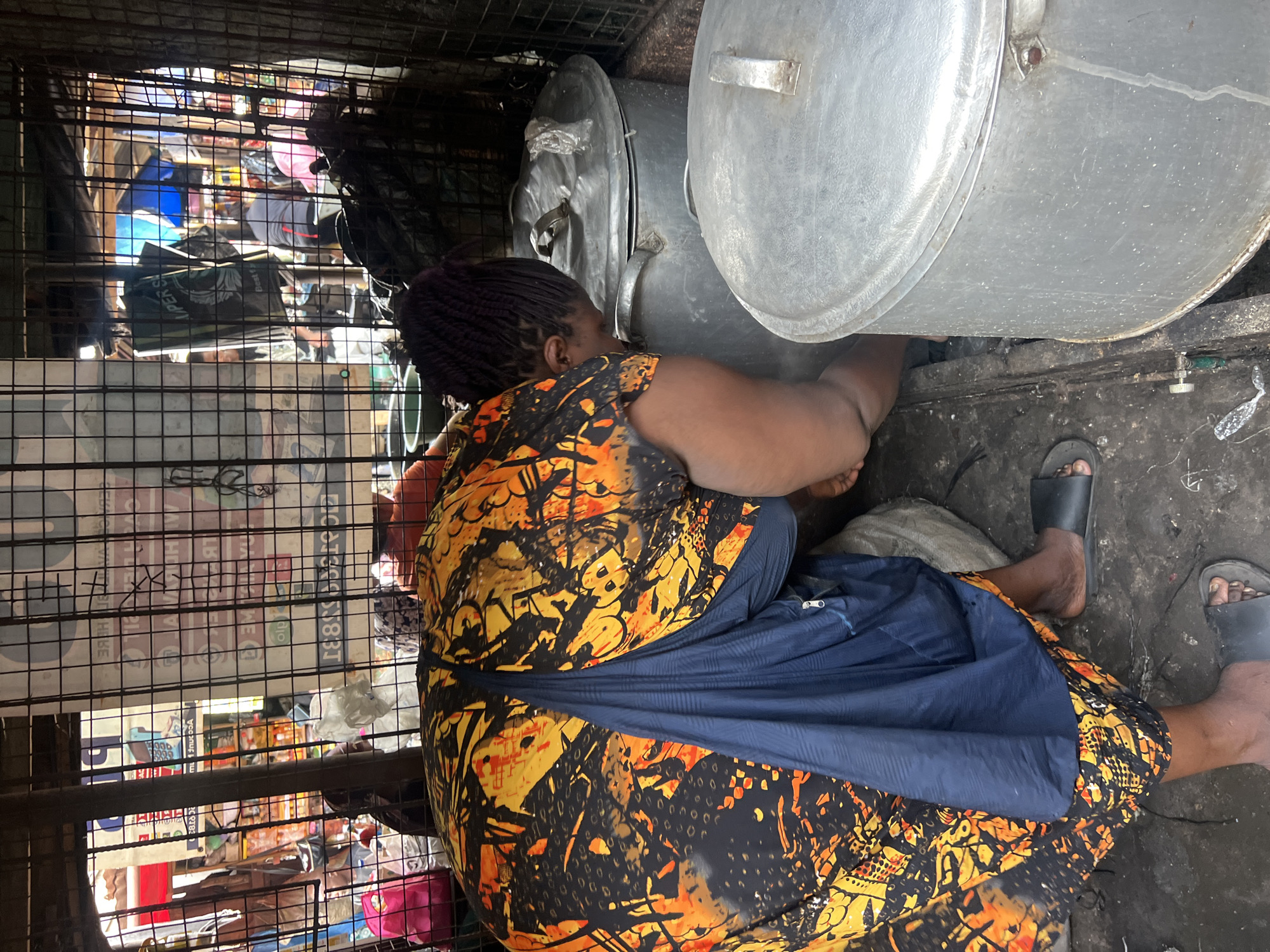 A woman in a patterned orange and black dress and a blue skirt is crouched between large metal pots and a metal cage. She wears black sandals. The area appears to be a small, enclosed space. The cage obscures the view of items in the background. The location is in the city of Unknown, Unknown.