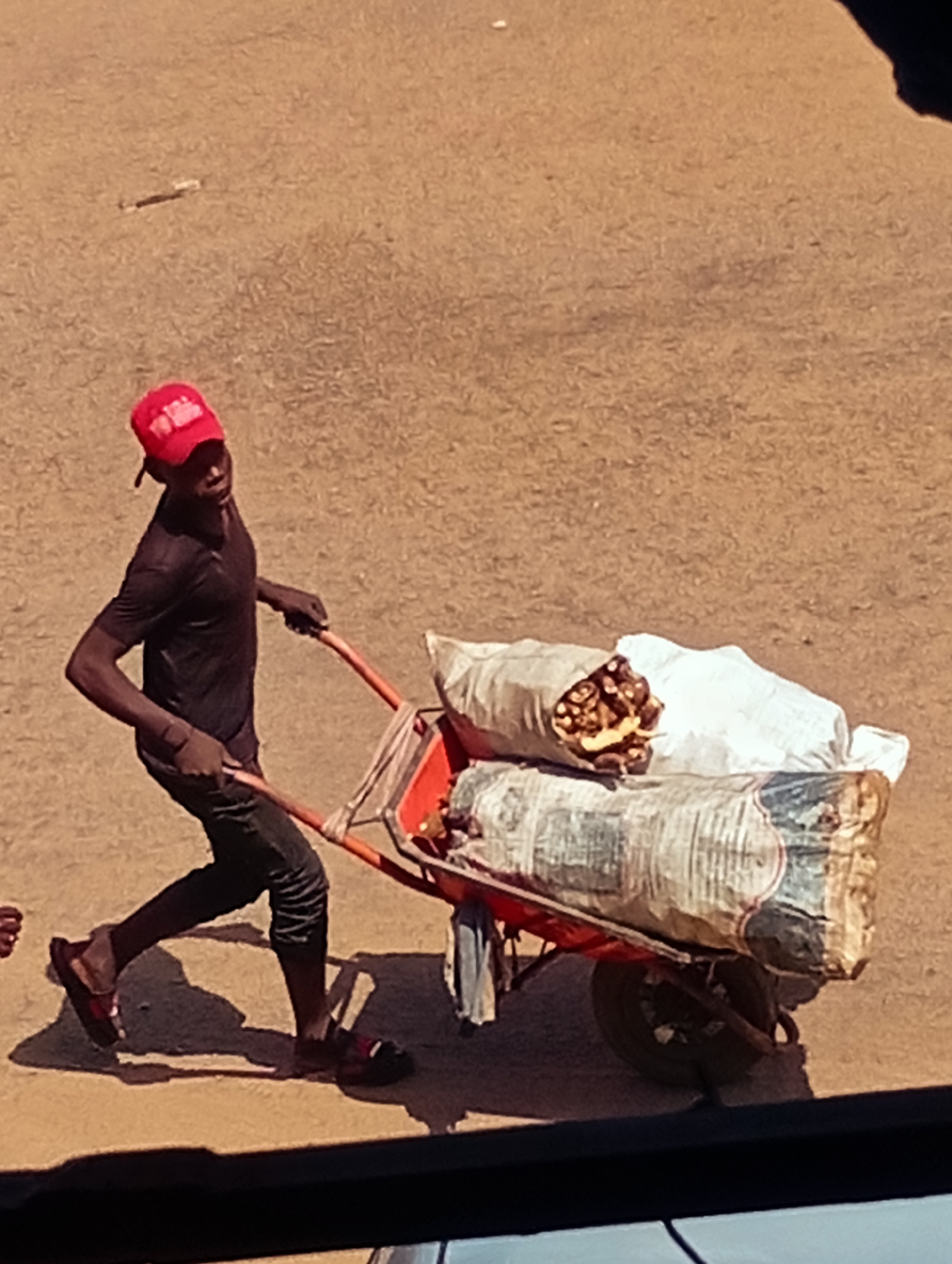 In Bukuru, Nigeria, a young person wearing a red baseball cap and a dark t-shirt pushes a two-wheeled cart laden with two large sacks. The cart's frame is orange, and one sack appears to be a light-colored, possibly burlap material, with some contents visible that resemble dried roots or produce. The second sack beneath is larger and darker, with some banding and labels that are difficult to decipher. The person is walking on a dirt or gravel surface, casting a distinct shadow to their left. The background is a uniform, dusty earth tone.