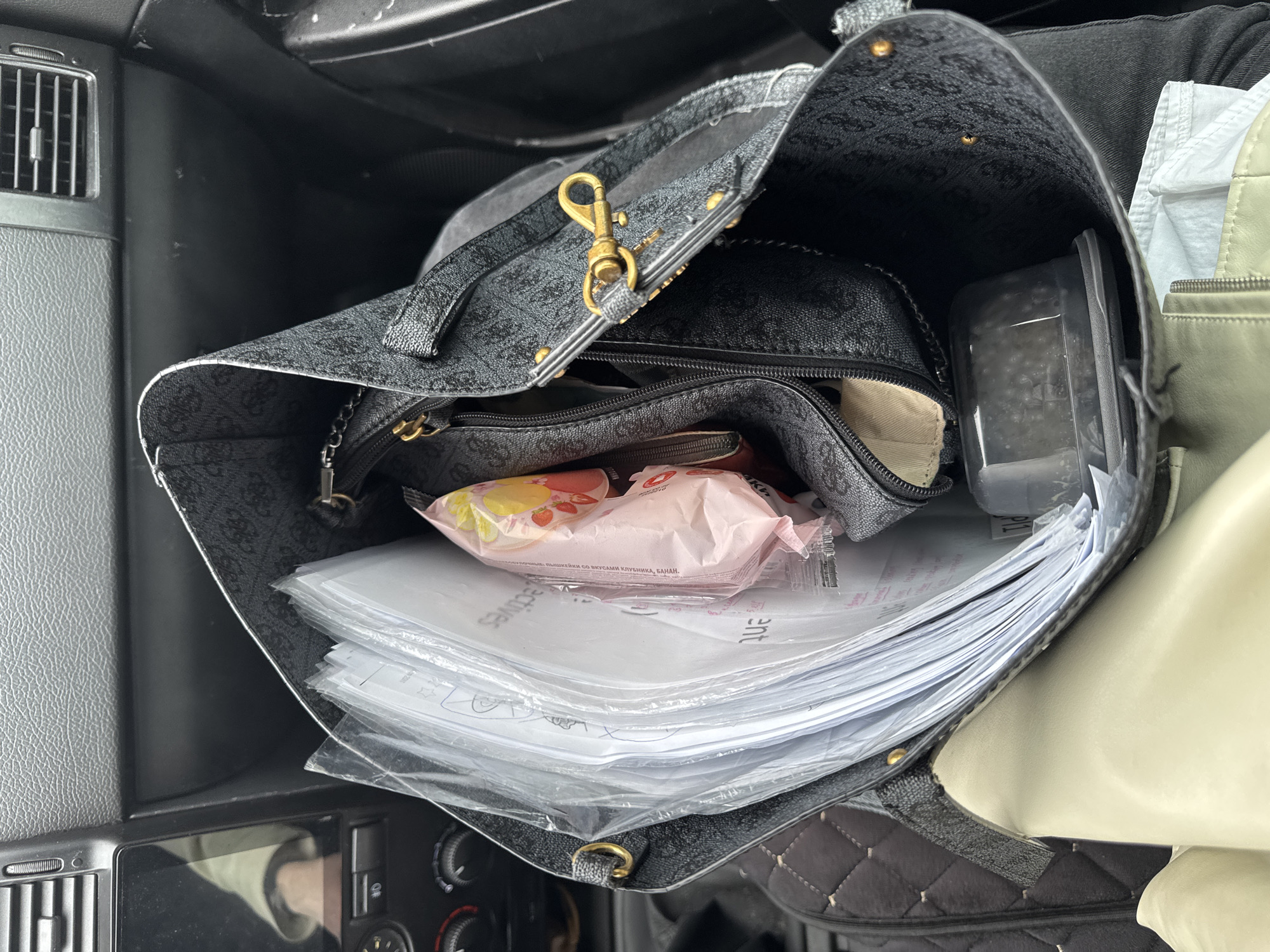 The image shows a gray patterned tote bag, seemingly placed within a vehicle. The bag appears to be open and filled with various items, including a stack of papers in plastic sleeves, a pink and yellow wrapped snack item, a zippered pouch, and a clear plastic container. The bag itself is sitting near a vehicle dashboard area featuring vents and control dials. The location in the city of Perm, Russia is not clearly indicated.