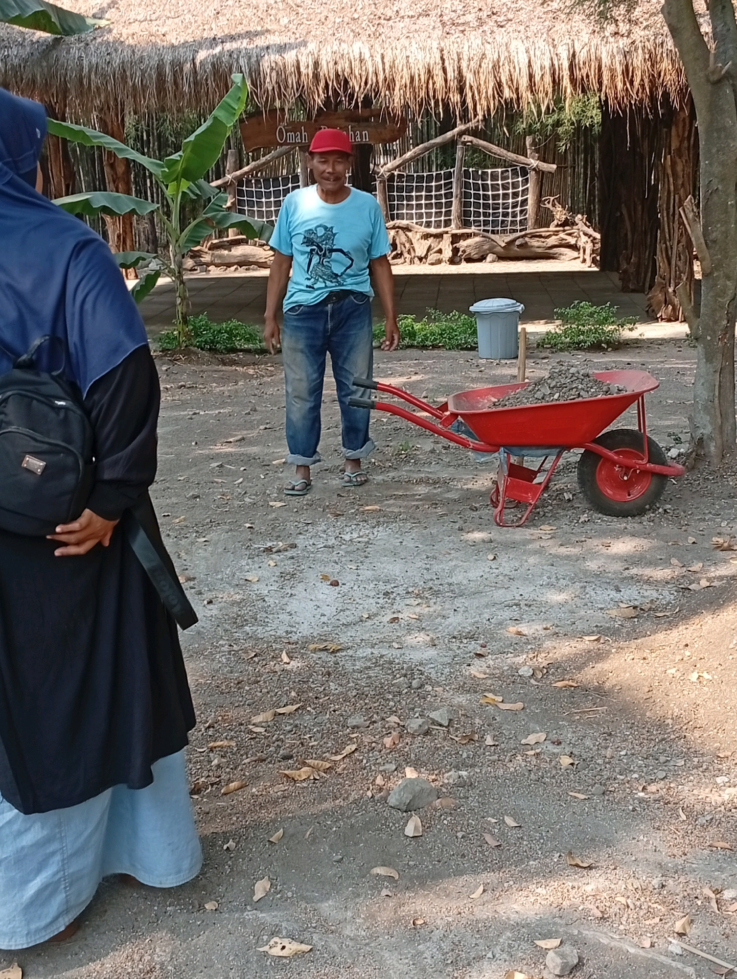 In Pandaan, Indonesia, a man wearing a blue t-shirt, blue jeans, and a red cap stands near a red wheelbarrow filled with gravel. He is positioned in the center of the frame, facing the viewer. To his left, a person wearing a dark blue hijab and a black outfit with a black backpack is partially visible. The environment appears to be an outdoor area with a thatched roof structure in the background, possibly part of a facility or attraction. Banana trees and dry leaves are scattered on the ground. A grey trash bin is also present in the scene. The lighting suggests it is daytime.