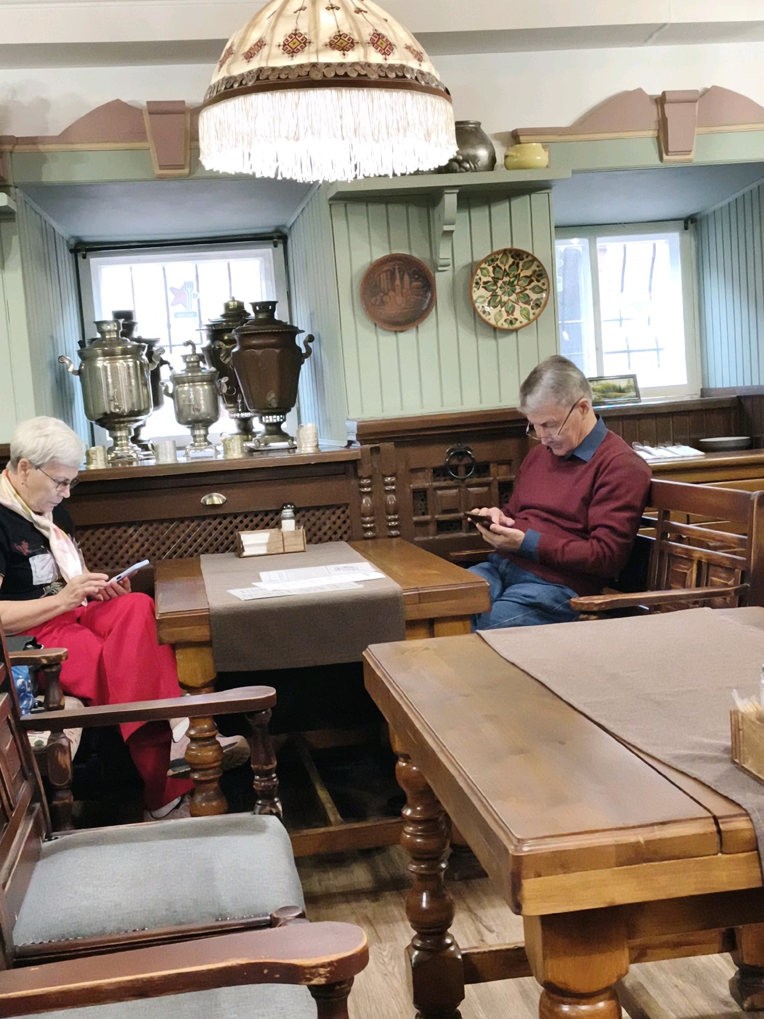 The image shows two individuals seated at tables in an indoor setting, possibly a cafe or restaurant, with a traditional Russian ambiance. On the left, a woman with short gray hair and glasses is wearing a black top and a red skirt. She is looking down at her mobile phone, which she holds in her hands. A light pink scarf is draped around her neck. She is seated in a wooden armchair with gray upholstery.
To the right, a man with gray hair and glasses is wearing a maroon sweater over a dark blue collared shirt and blue jeans. He is also looking down at a mobile phone in his hands. He is seated on a wooden bench.
The background features wooden furniture, including a dark wooden cabinet with decorative carvings. Several samovars are displayed on a shelf above the cabinet, suggesting a cultural or historical theme. Decorative plates hang on the light green walls. Two windows are visible, allowing natural light into the space. The tables are covered with brown tablecloths.
The scene appears to be in Cheboksary, Russia, given the presence of samovars and the overall traditional aesthetic, which is characteristic of some establishments in the city.