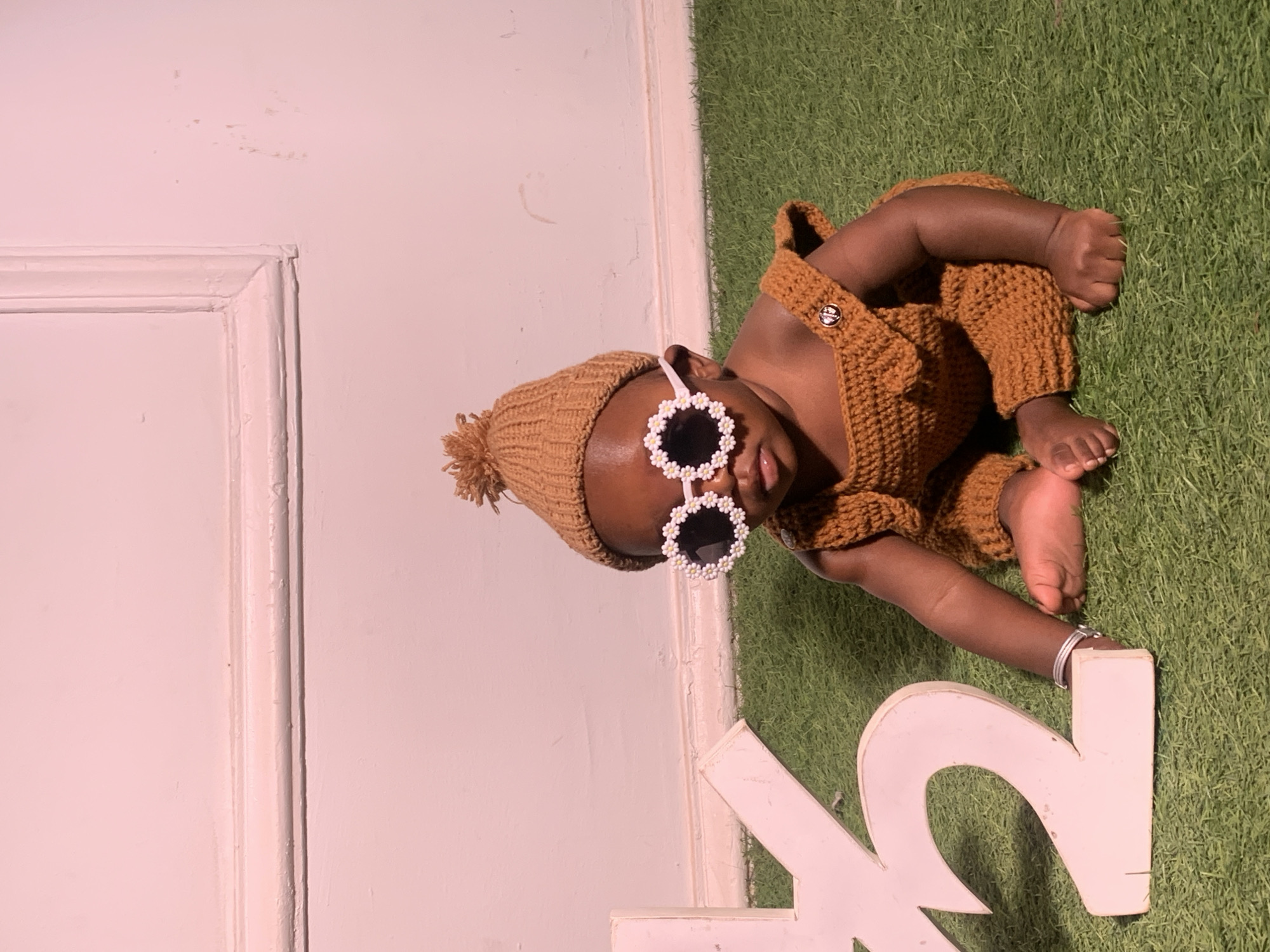 A baby lies on an artificial turf background adjacent to a white wall. The baby wears a brown knitted hat and matching overalls. Flower-shaped sunglasses are positioned on the baby's face. A white decorative "K" is near the baby's feet. The location is presumed to be a studio or indoor setting; there is no evidence of Barakin Akawo, Nigeria.