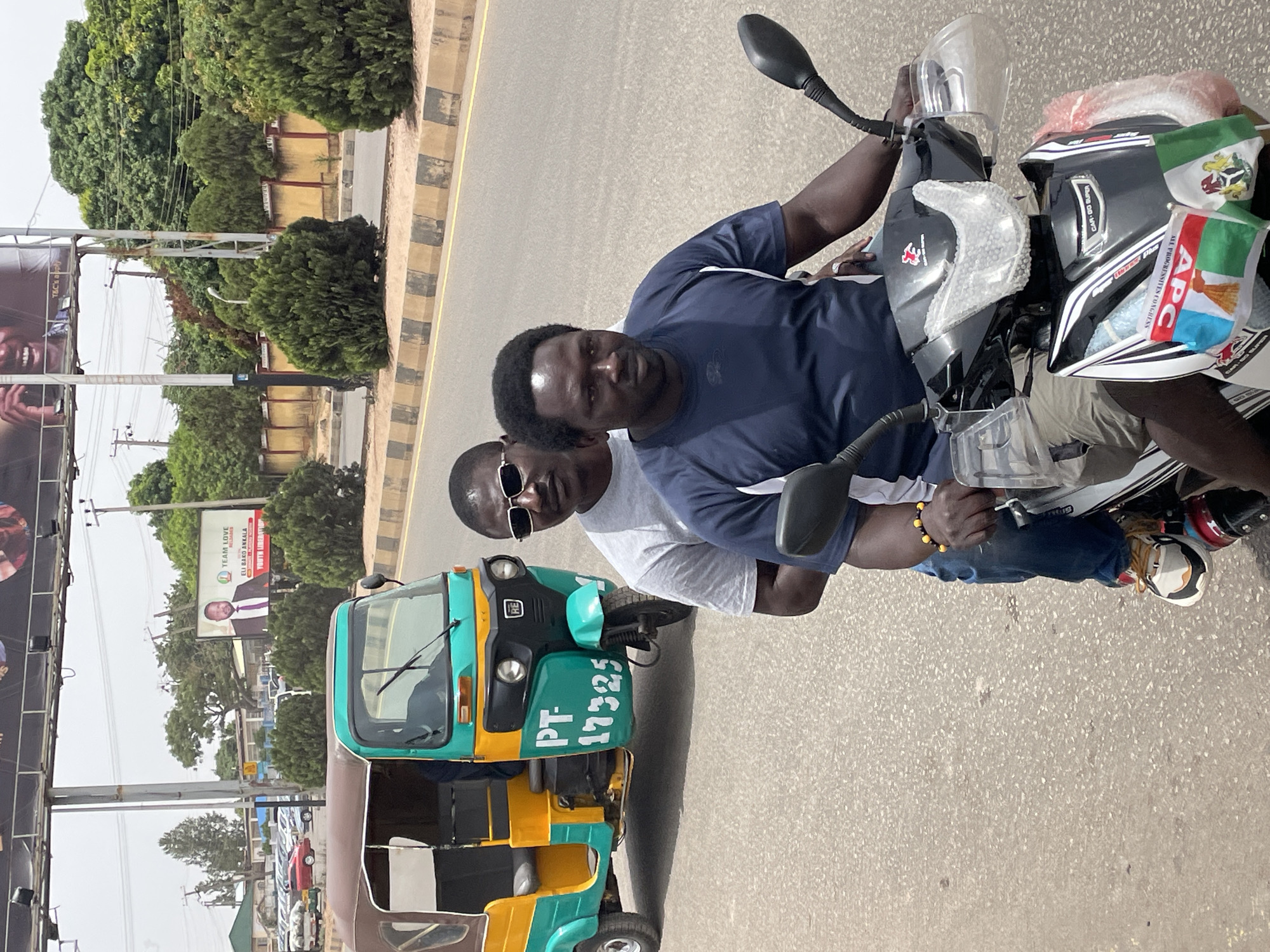 An outdoor street scene in Jos, Nigeria, captures a snapshot of daily life. The image, taken from a slightly elevated perspective and rotated ninety degrees clockwise, centers on two men on a black and silver motorcycle. The rider, wearing a dark blue t-shirt, looks directly at the camera, while his passenger, in a white t-shirt and sunglasses, is partially obscured behind him. The motorcycle's front is adorned with small flags, including the Nigerian national flag and one for the "APC" (All Progressives Congress) political party, also displaying "ALL PROGRESSIVES CONGRESS" and "CAR-DO SUPER".
To the left of the motorcycle, a yellow and green three-wheeled vehicle, commonly known as a Keke Napep or tricycle, is visible. The license plate or identification number "PT-17325" is prominently displayed on its front. Both vehicles are on an asphalt road with distinct black and yellow striped curbs, suggesting a main thoroughfare.
In the background, a bustling street environment unfolds. Lush green trees and bushes line the road. Several billboards are visible; one on the far left has "T&C's apply" partially legible, while another further down features a man's portrait and text that reads "TEAM LOVE", "ELI BAKO ANKALA", and "YOUTH LIBERATIO", indicating political or community promotion. Other vehicles are parked in the distance. The bright lighting suggests it is daytime, likely a clear and sunny day.
