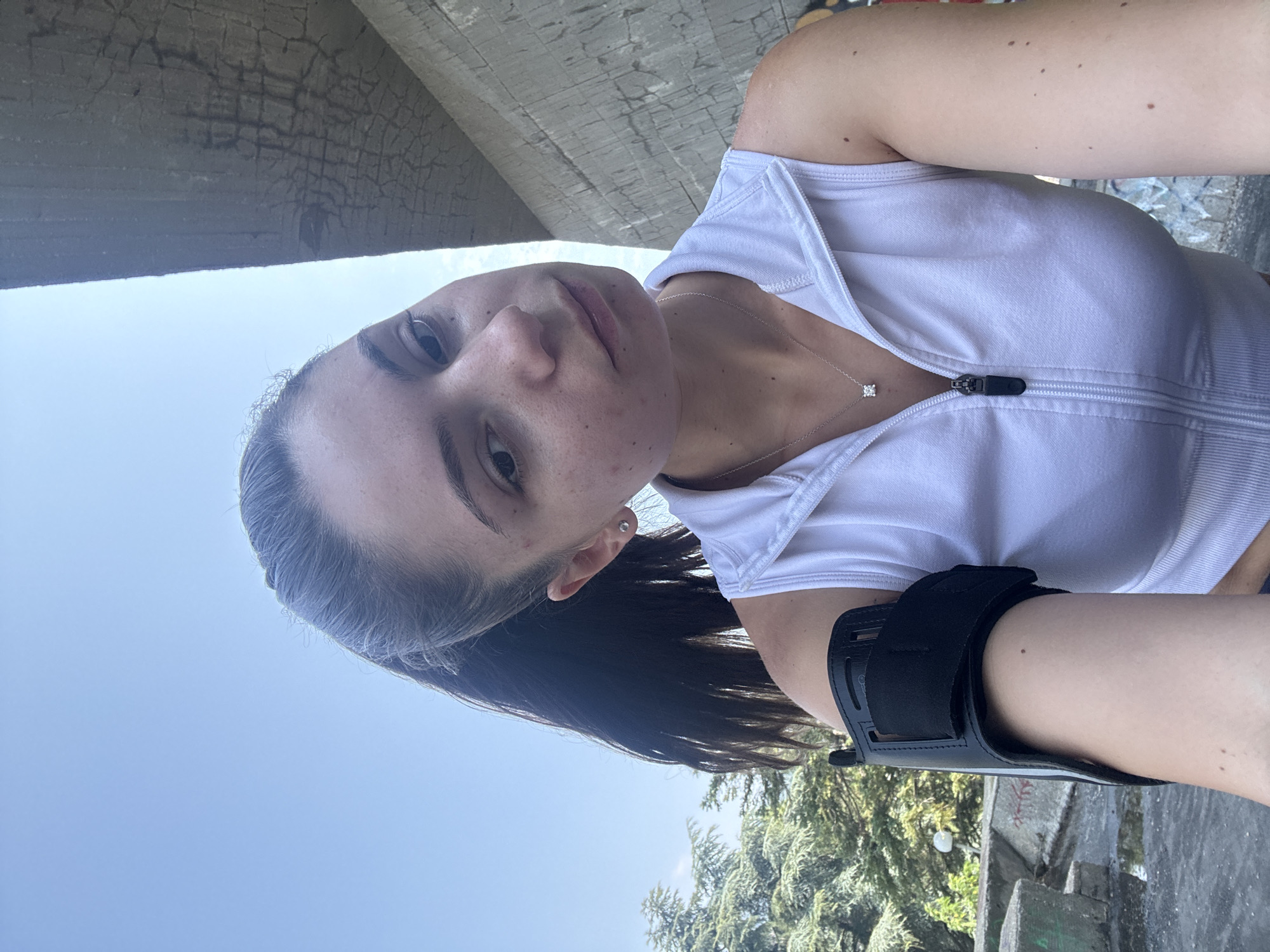The media file shows a young woman in a white tank top and a black arm brace. She has dark hair pulled back and is looking at the camera. Her face is tilted slightly, and she has a faint smile. She is wearing a silver necklace with a small pendant. The background suggests an outdoor setting with a bright blue sky and trees. There is also a concrete structure visible, possibly a bridge or underpass, with a rough, weathered surface. The lighting indicates it is daytime, likely sunny. The overall impression is casual and possibly after some physical activity, given the arm brace. There are no visible text elements in the image.