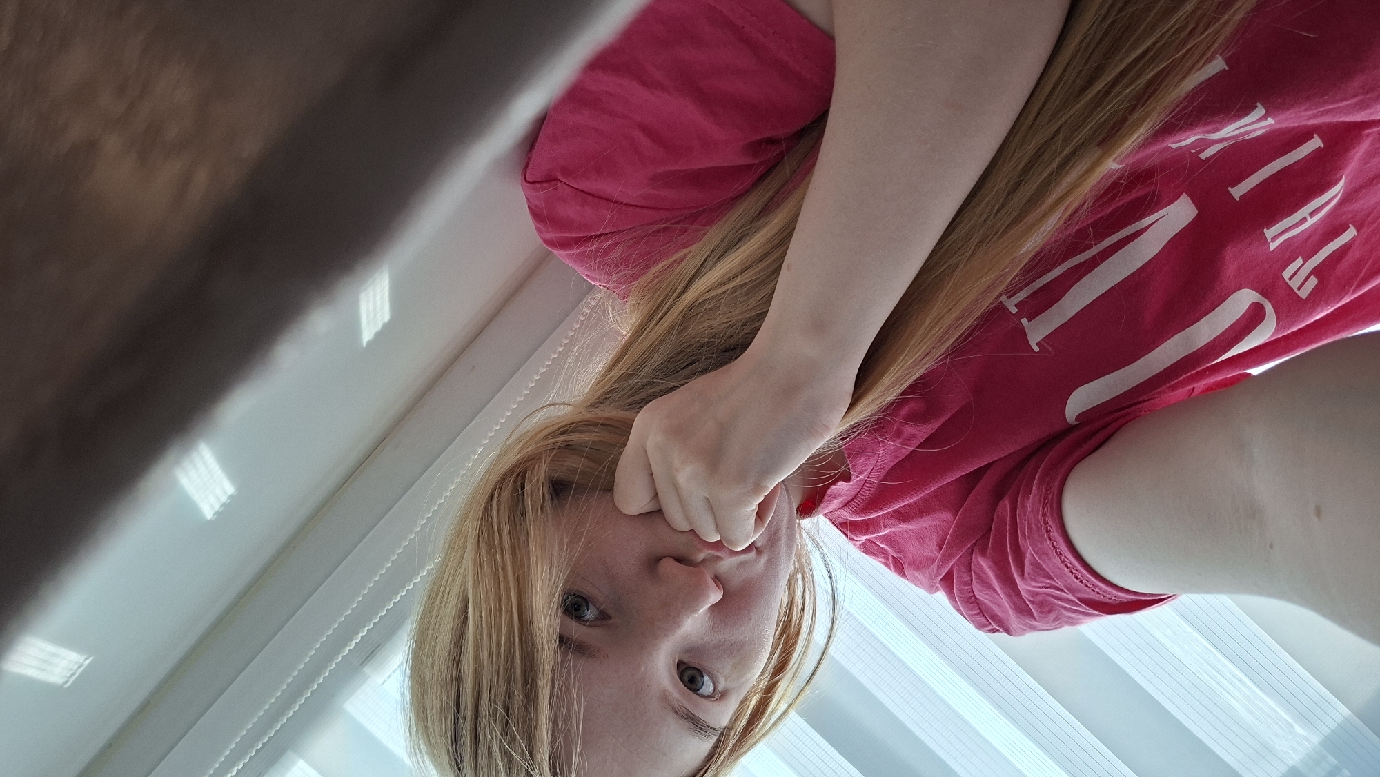 A woman with blonde hair and blue eyes is looking directly at the camera. She is wearing a pink t-shirt and has her right hand touching her face near her mouth. The background includes a window with white blinds and a portion of a wall. The location is Chaikovsky, Russia.