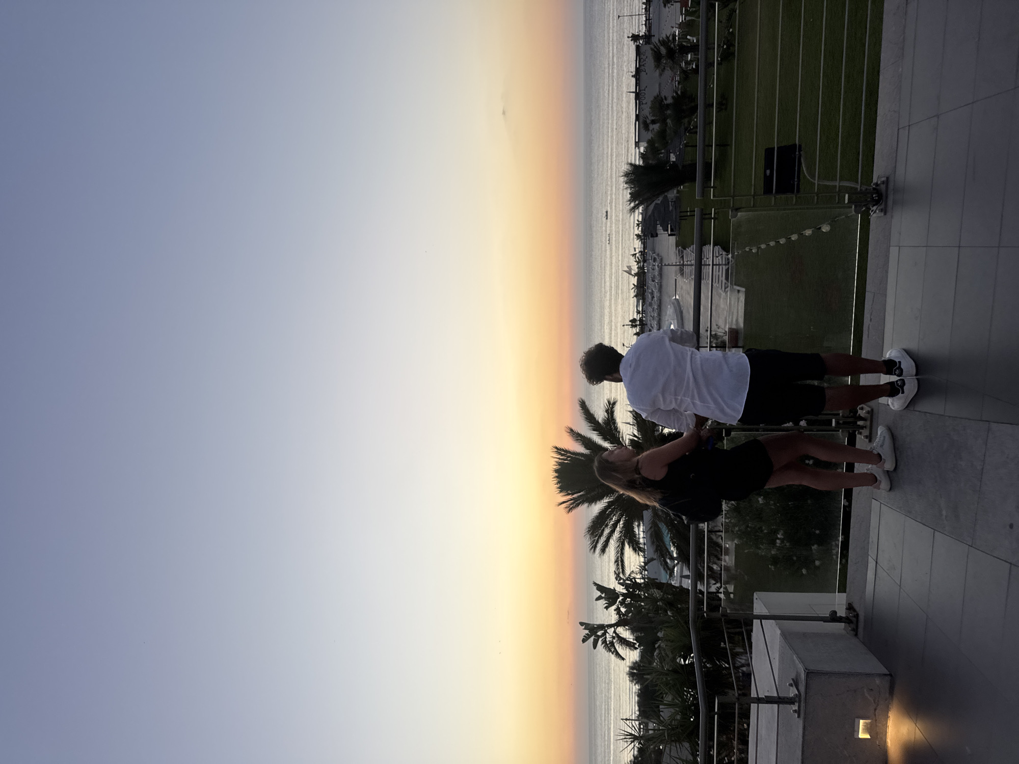 Two individuals, a woman and a man, stand on a tiled balcony, looking out at the sea during sunset. The woman, on the left, wears a black dress and carries a backpack. The man, on the right, is dressed in a white shirt and black shorts. In the background, palm trees and other vegetation are visible, along with a beach, some lounge chairs, and structures suggesting a resort or hotel. The sky displays a gradient of colors, transitioning from light blue at the top to orange and pink near the horizon. The sea is calm with a few small boats visible in the distance. The scene appears to be located in Isola delle Femmine, Italy.