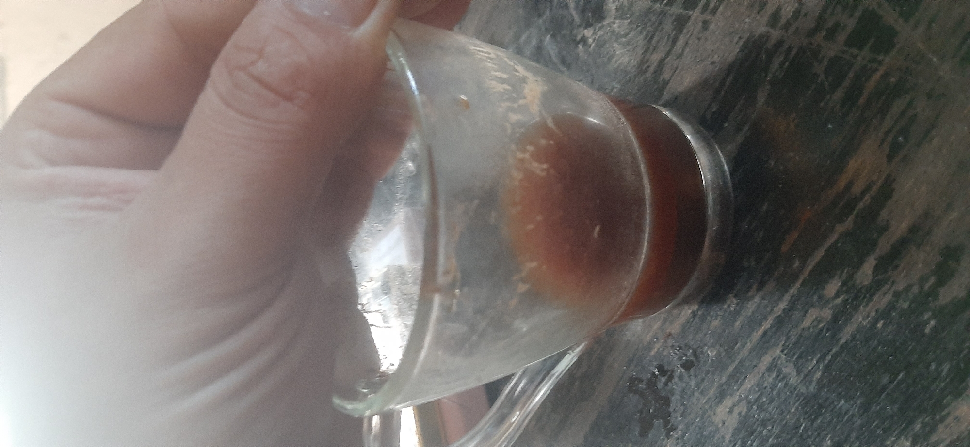 A close-up, overhead shot shows a hand with visible knuckles and fingernails, partially obscuring the top of a clear glass mug filled with a dark liquid, likely coffee. The mug sits on a dark, weathered wooden surface with a visible grain and streaks of lighter and darker wood. Some dark liquid appears to have spilled onto the surface next to the mug. The background is blurred and appears to be an indoor setting, possibly a table. The image was taken in Yessentukskaya, Russia.