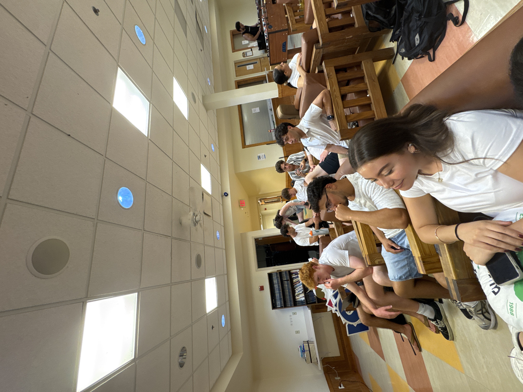 The image shows a group of young adults gathered in a common room, possibly a lounge or recreational area. They are seated on wooden chairs and sofas, and appear to be watching something. Some individuals are looking towards the front of the room, while others are engaged in conversation or looking at their phones.
The room has a tiled ceiling with recessed lighting and ceiling fans. The walls are painted a light color, and there are wooden accents. In the background, there is a bookshelf filled with books, a desk with some items on it, and an exit sign. A foosball table is visible in the far background. The floor is tiled with a geometric pattern.
The people are casually dressed in t-shirts and shorts. Their ages appear to be in the late teens to early twenties. The overall atmosphere suggests a relaxed social gathering. The location is identified as Washington Township, United States of America.