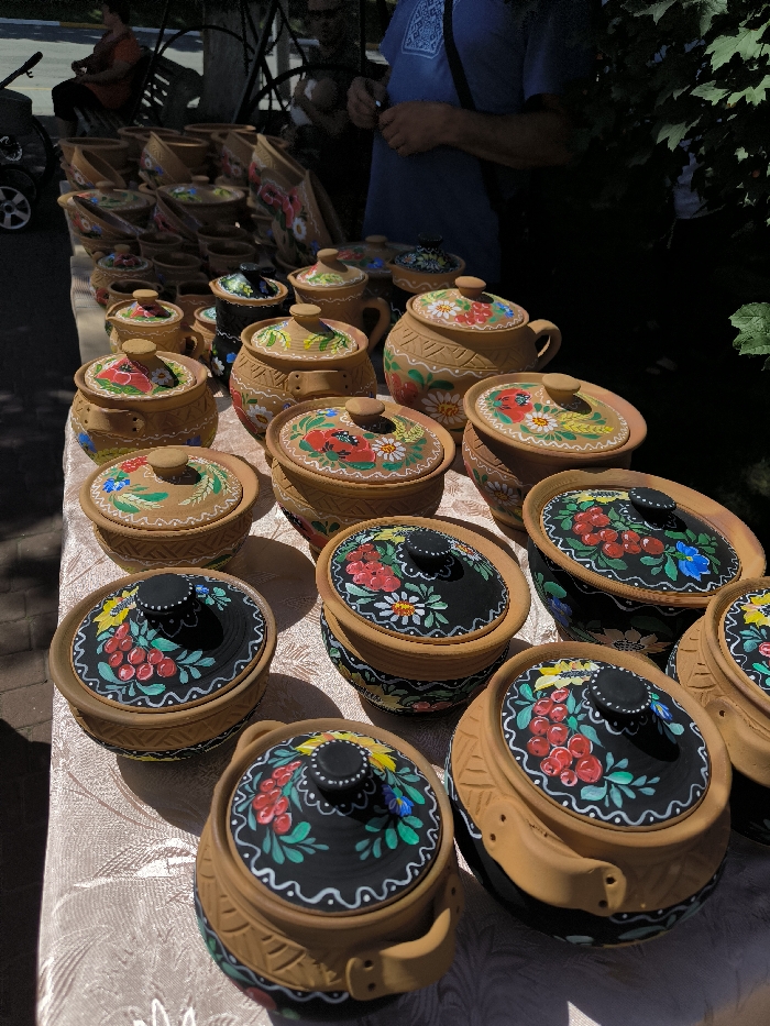 The image displays an outdoor market stall in Illintsi, Ukraine, showcasing approximately thirty pieces of traditional Ukrainian earthenware pottery under clear daylight conditions. Numerous lidded pots and bowls, crafted from reddish-brown terracotta, are meticulously arranged on a light-colored, draped table. Each pottery item is hand-painted with intricate floral and botanical motifs; prominent designs include clusters of red berries (likely kalyna), green leaves, and various white, yellow, and blue flowers. Several lids feature a contrasting black background for these painted designs, while others are decorated directly onto the natural clay surface, often complemented by geometric patterns on the pot bodies.
In the background, a male individual wearing a blue t-shirt with a light embroidered pattern is partially visible standing behind the stall. Further left, another person is partially visible, seated on a bench or similar structure. The outdoor environment is characterized by strong natural sunlight casting distinct shadows, green foliage on the right side, and a paved surface visible on the far left.