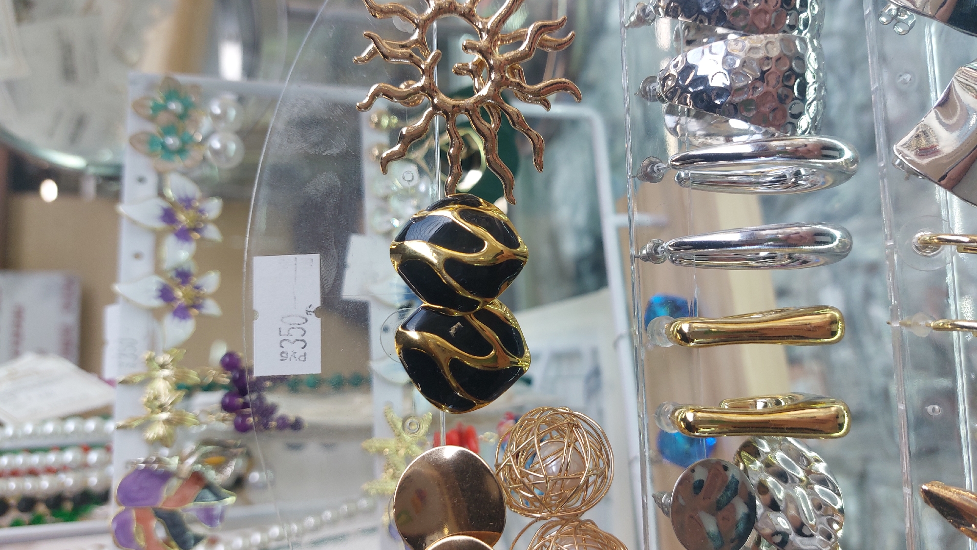The image captures a selection of earrings displayed in a store window, presumably in Novosibirsk, Russia, as indicated by the price tag "PYб" (likely an abbreviation for Russian Rubles).
Several types of earrings are visible. The central focus is a pair of black and gold earrings with a sun-like ornament at the top. To the right, there's a rack of earrings with silver and gold designs. To the left, behind a semi-transparent display, are earrings with floral and other decorative elements. A price tag with "3350" is also visible near those earrings.
The earrings are displayed on clear plastic racks, which are typical for jewelry store displays. The background is blurred, suggesting the store's interior and possibly other merchandise.