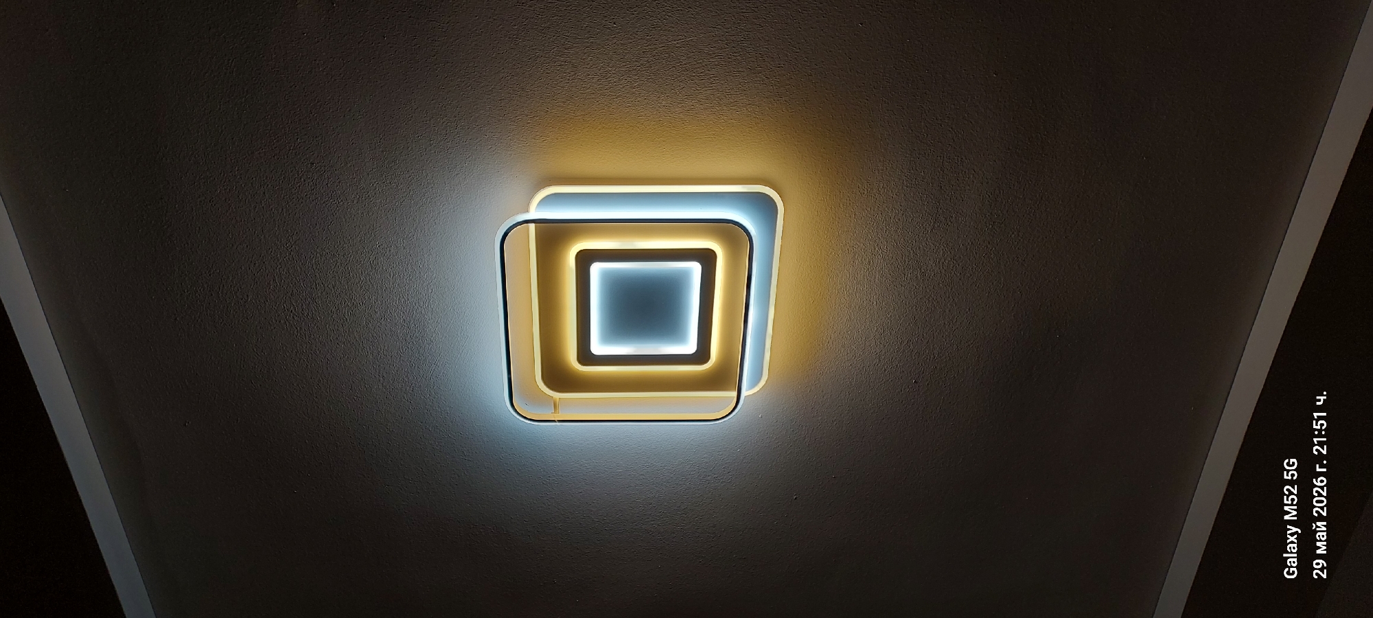 A ceiling-mounted light fixture is the central focus of the image. The fixture is a square design with concentric squares of white and yellow light. The surrounding ceiling is off-white with a textured surface. There is a black border visible on the left and right edges of the image. The date and time of the photo, "29 май 2026 г. 21:51 ч.", is embedded at the bottom right corner. The photo was taken with a Galaxy M52 5G. There is no evidence of people or the city of Canals, Spain.