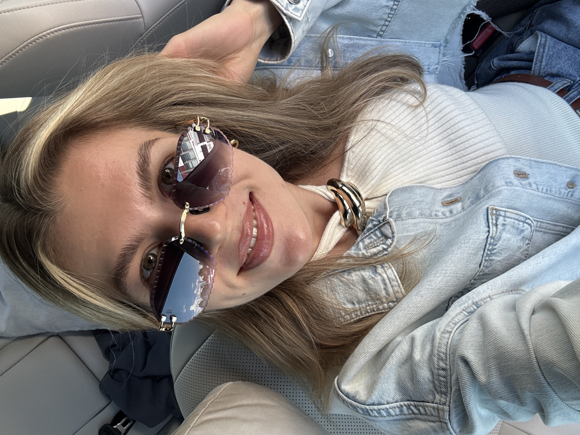 A blonde woman, wearing large, stylish sunglasses and a denim jacket over a white ribbed top, smiles at the camera from what appears to be the passenger seat of a car. The interior of the car is visible, with a grey seat and headrest. Her hair is partially obscuring her face, and she is touching her hair with her right hand. The sunglasses reflect the sky and buildings, suggesting an outdoor, possibly urban, setting. A dark blue garment is visible on the seat behind her, and part of a seatbelt is also visible. The overall impression is a casual, candid shot taken during the daytime.