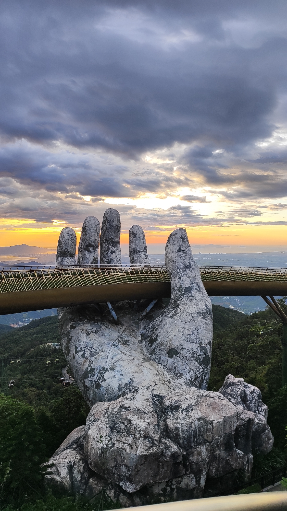 This image captures the Golden Bridge in the Ba Na Hills of Da Nang, Vietnam, at sunset. The iconic bridge is held aloft by two colossal stone hands, weathered and textured to resemble ancient rock formations. A golden railing with intricate metalwork lines the walkway of the bridge, which stretches between the fingers of the left hand and disappears behind the right. The bridge spans a lush, green mountain landscape dotted with cable cars, suggesting a resort or park setting. In the background, a hazy city skyline and distant mountains are visible against a dramatic sky painted with hues of orange, yellow, and purple, indicative of sunset. The clouds are heavy and dramatic, with patches of light breaking through, adding to the atmospheric quality of the scene. The composition emphasizes the scale and unique design of the Golden Bridge against the natural beauty of the surrounding environment.
