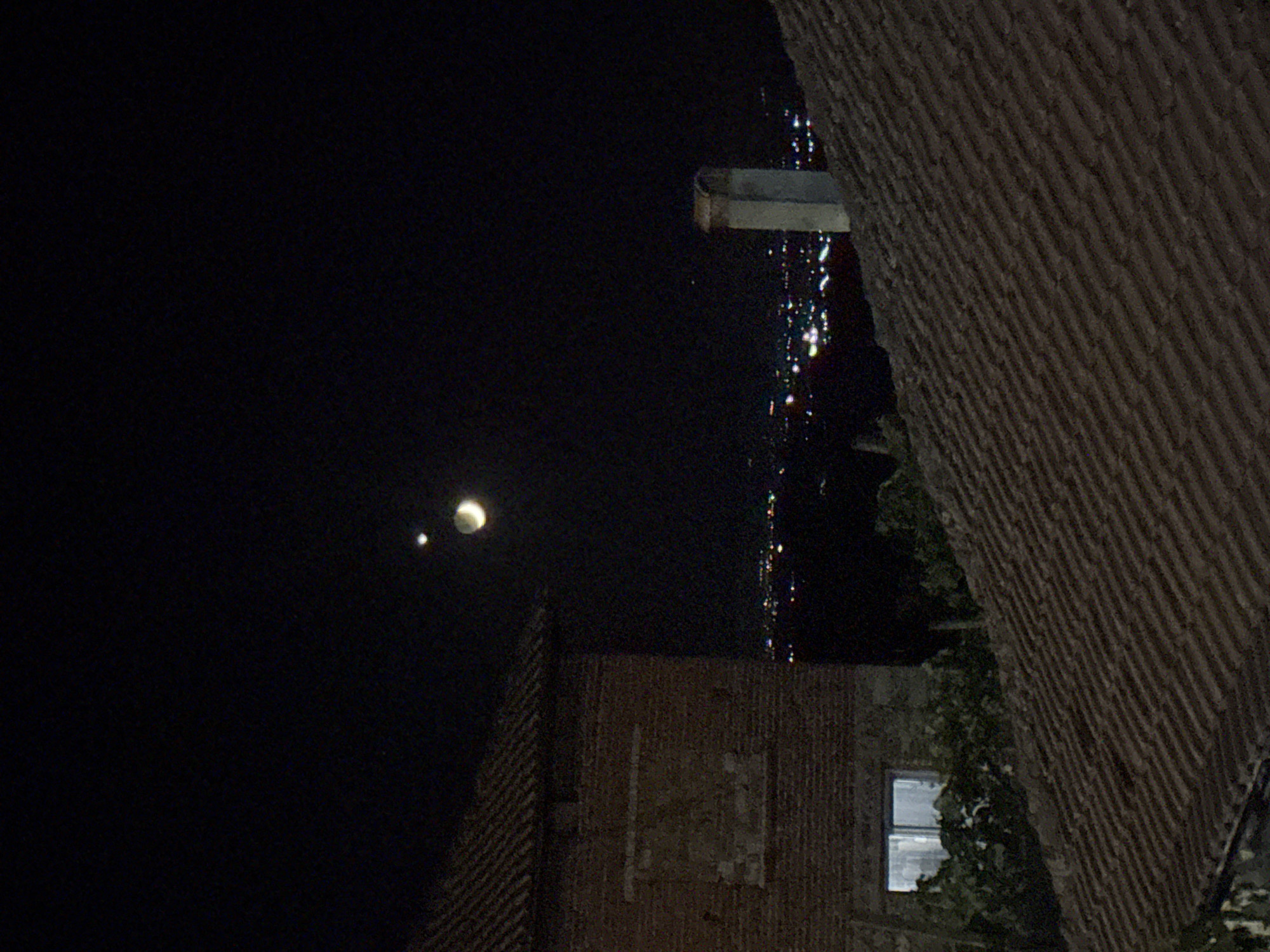 A nighttime scene captures a view over a city's rooftops. In the foreground, a dark, tiled roof slopes downwards, partially obscuring a brick building with a large window. Above the distant city lights, the crescent moon is visible in the dark sky, accompanied by a small, bright point of light, likely a planet or star. A chimney with a rounded top stands on another rooftop in the middle ground, silhouetted against the illuminated cityscape. The city of Skrat, Bulgaria is visible in the background.