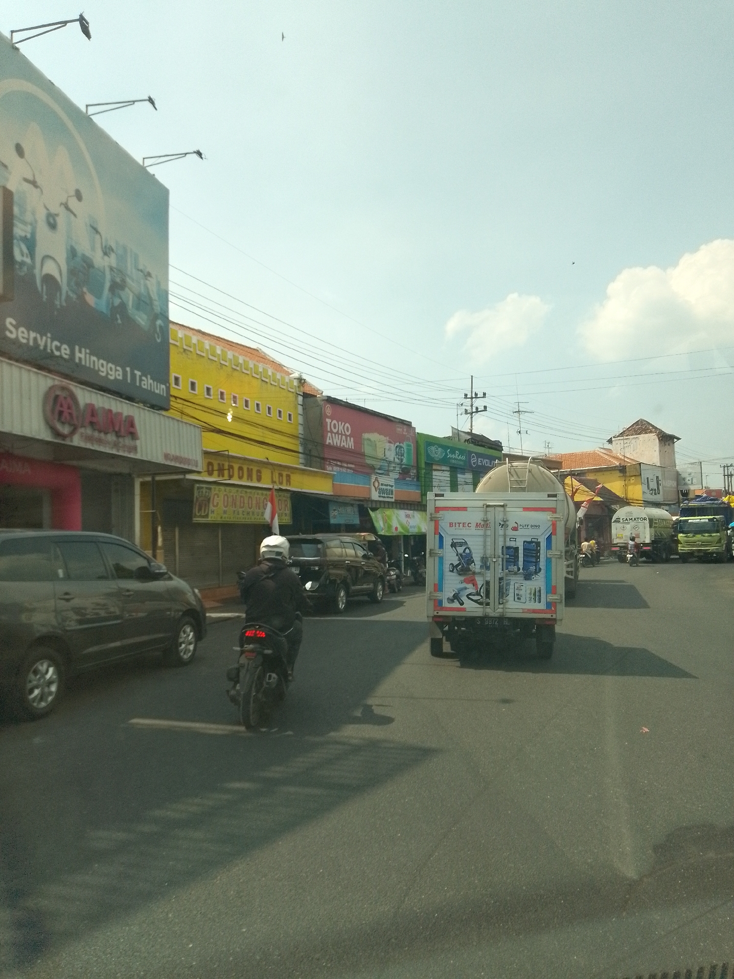 The image captures a street scene in Babat, Indonesia, under a bright, partly cloudy sky. The perspective is from a vehicle moving along a paved road. On the left, a dark-colored SUV is parked. Further left, a large billboard with an advertisement for "Service Hingga 1 Tahun" (Service Up to 1 Year) and an image of scooters is visible. Adjacent to the billboard is a shop with a red awning and the brand name "YAMAHA".
Across the street, a row of buildings displays various shopfronts and advertisements. Notable among them are shops with yellow facades and signs reading "ONDONG LOR" and "TOKO AWAM" (Awam Store). Red and white Indonesian flags are displayed on some buildings.
The road is populated with vehicles. A person on a motorcycle wearing a helmet is in the foreground, moving away from the viewer. Behind the motorcycle, a black SUV is driving in the same direction. In the distance, several trucks are on the road. One truck, prominently in the center, is a white box truck with advertisements on its rear, including "BITEC MOT" and "PUFF DINO". Another truck is a tanker truck with "SAMATOR" written on its side, and a green truck is further down the road. Power lines and utility poles are visible overhead, typical of urban infrastructure. The overall impression is a bustling commercial street with ongoing traffic.