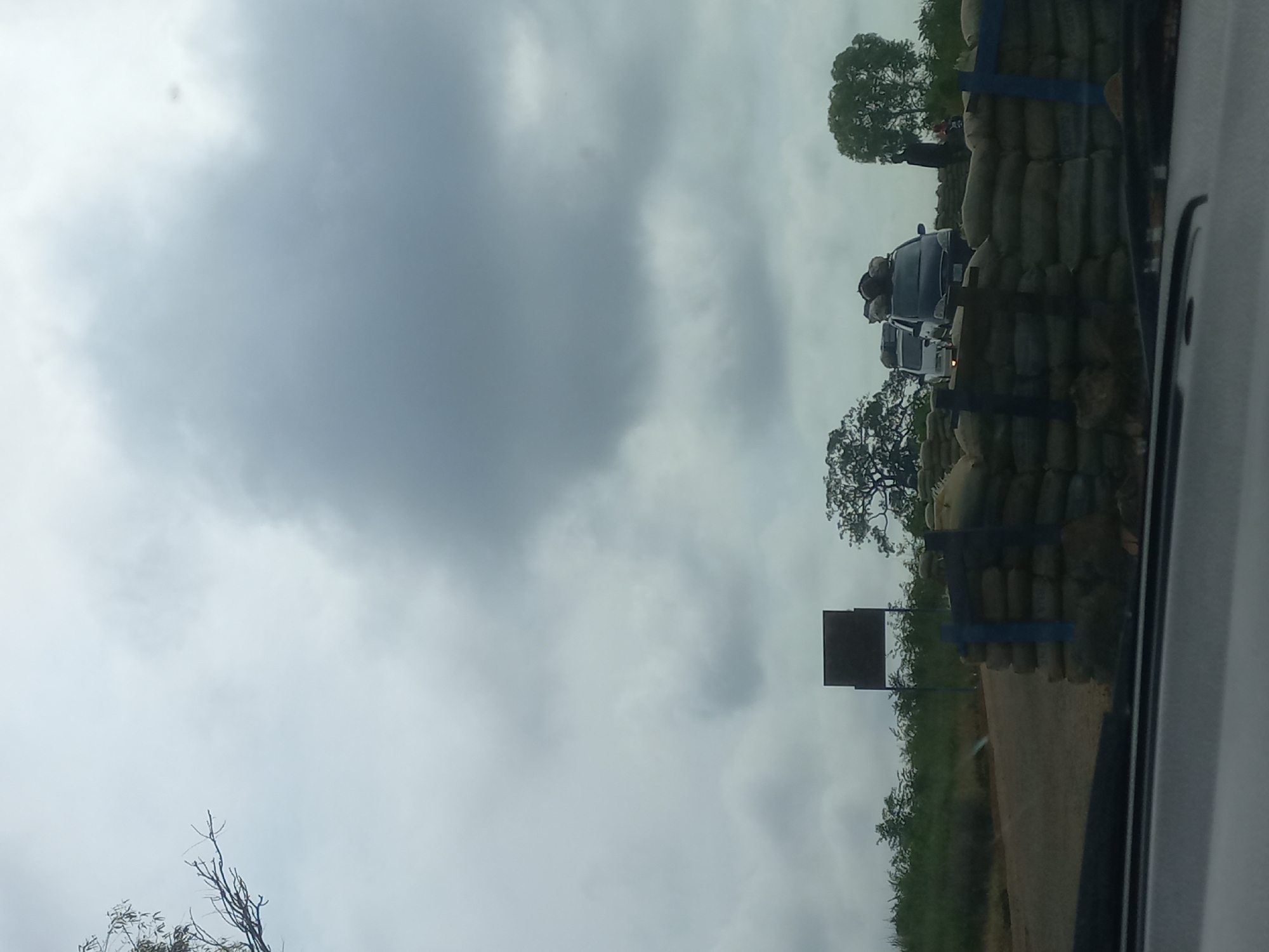 In Mangu Halle, Nigeria, a dark, overcast sky looms over a scene of vehicles queuing behind a barricade constructed of sandbags and blue posts. The barricade, positioned on the right side of a dirt road, appears to be a checkpoint or a security measure. Several vehicles are visible, including a dark SUV laden with luggage on its roof, and a white van. People are standing near the barricade and vehicles, some appearing to be in conversation or directing traffic. The surrounding environment consists of lush green vegetation and trees, with a dark, rectangular signpost visible in the distance. The foreground is dominated by the interior of a vehicle, suggesting the photograph was taken from inside a car. The overall impression is one of a temporary disruption or security presence on a rural road.