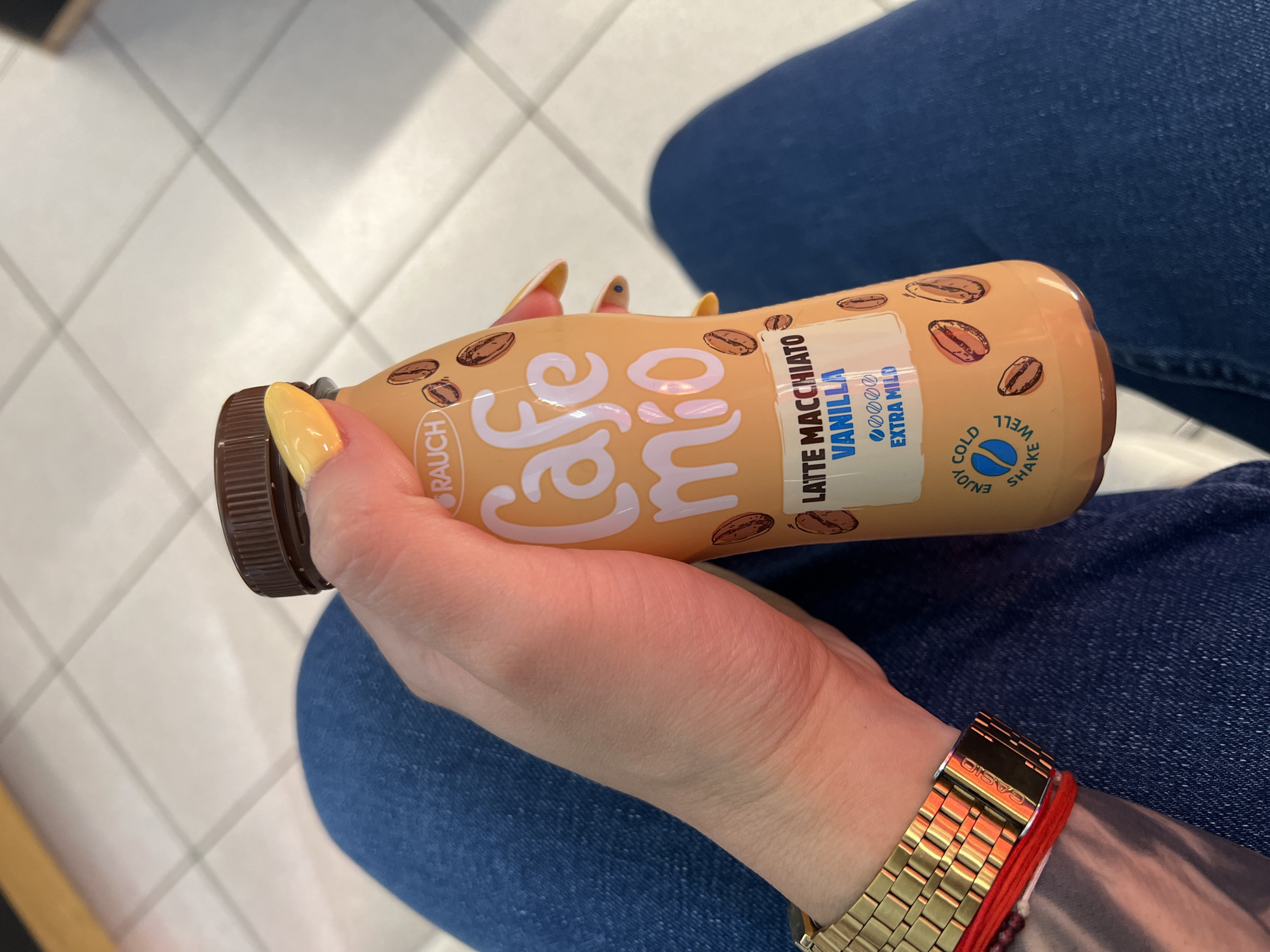 A person's hand, adorned with yellow nail polish and a gold Casio watch with a red string bracelet, holds an orange bottle of "Cafe mio Latte Macchiato Vanilla Extra Mild." The bottle has coffee bean graphics and text indicating "Enjoy Cold Shake Well." The background shows a tiled floor and blue denim fabric, suggesting the person is seated. The specific location in Pazardzhik, Bulgaria cannot be determined from the visual information.