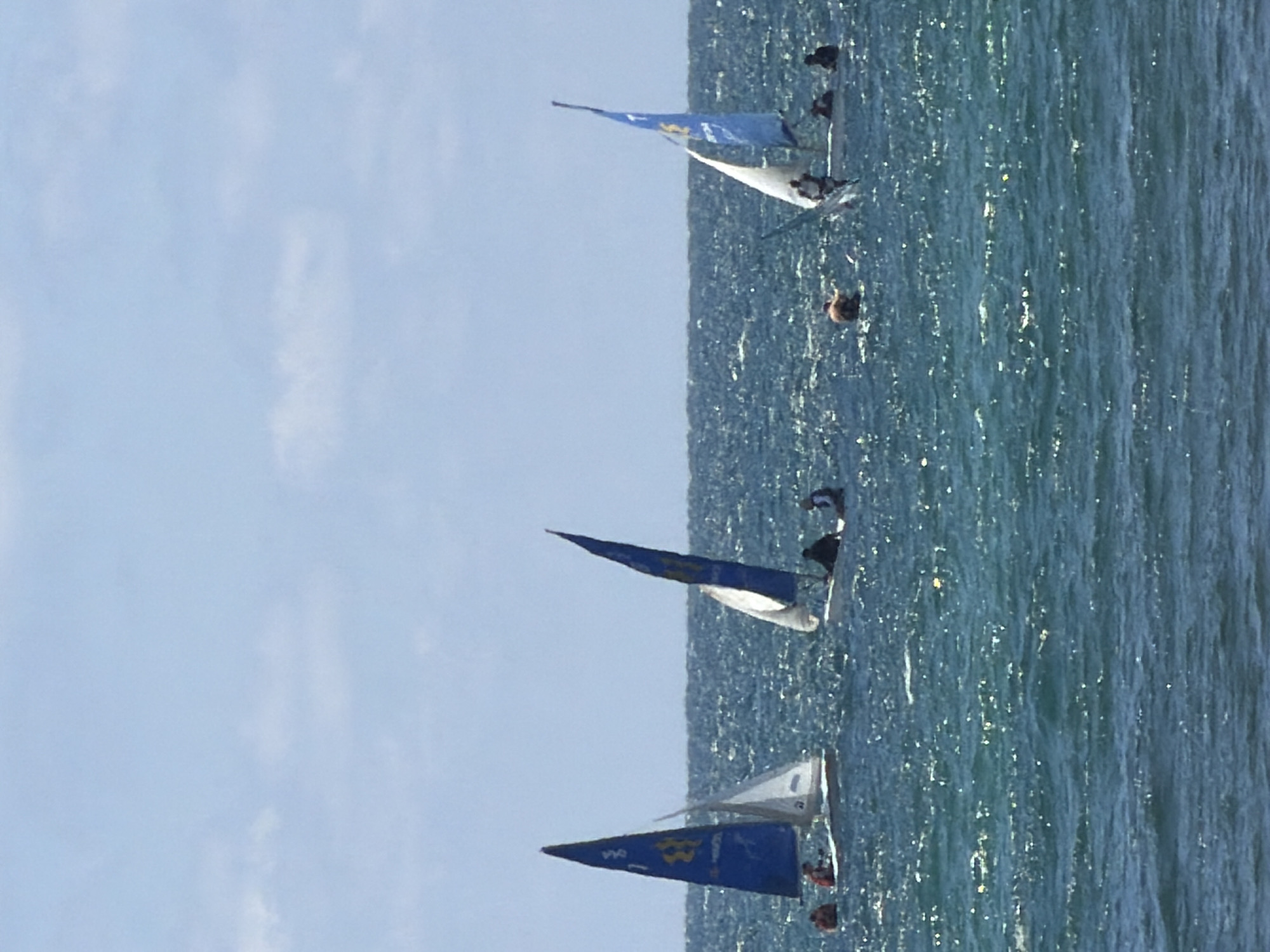 The image shows a seascape off the coast of Tel Aviv, Israel, featuring several small sailboats. The boats are equipped with blue and white sails and appear to be in a light breeze. The water is choppy with sunlight reflecting off the surface. Small figures that appear to be people are visible on the boats, seemingly engaged in sailing activity. The sky is light blue and mostly clear.