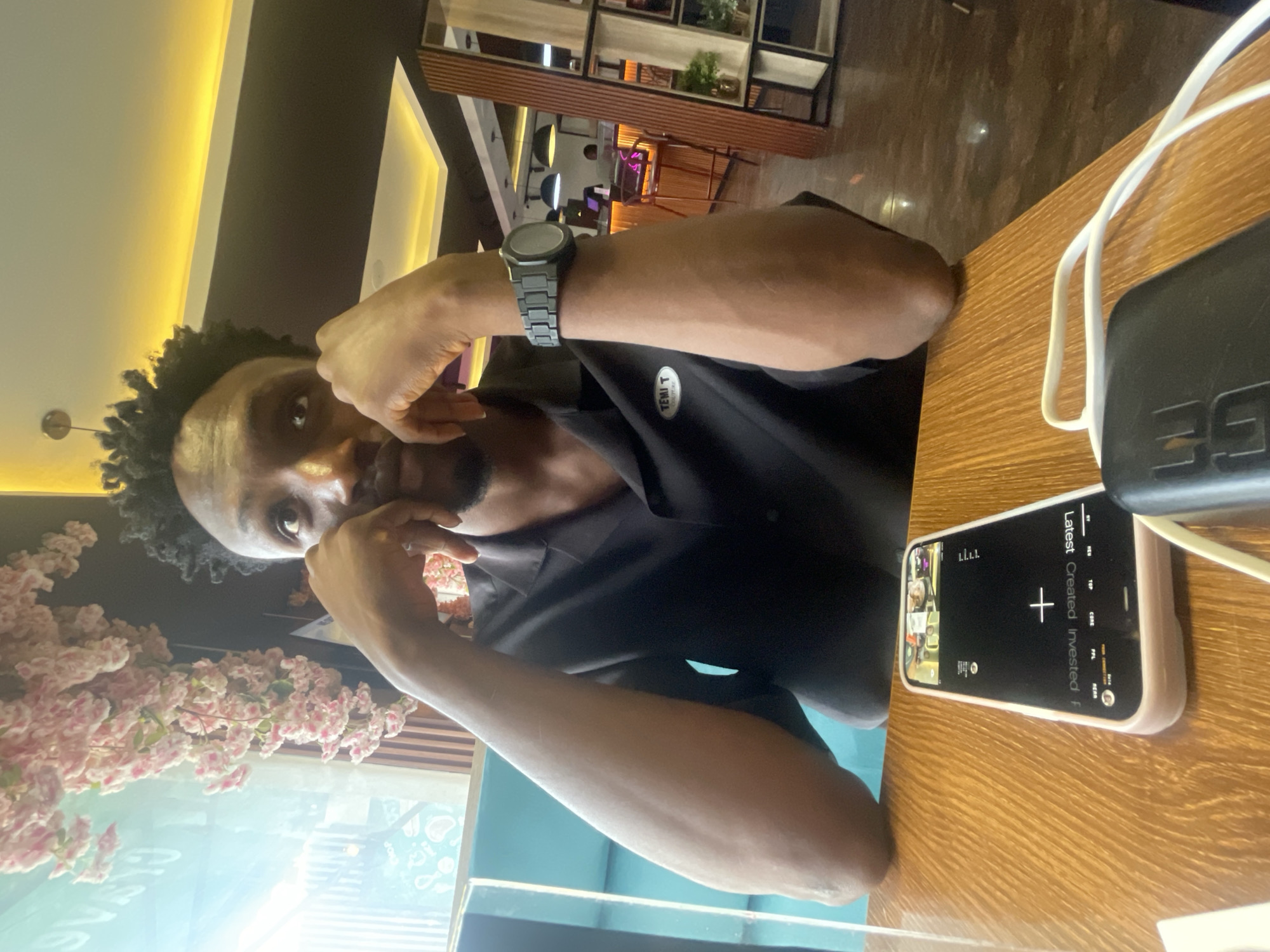 A man with short, dark hair is seated at a wooden table indoors. He is wearing a black shirt and a black smartwatch. His left hand is near his face, and his right arm is on the table. A phone with a blank screen is on the table to his right, along with a black object and a white cable. Through a window, possibly a glass barrier, there are pink flowers and a view of Adankolo, Nigeria. In the background are shelves and chairs. The location seems to be a restaurant or cafe.