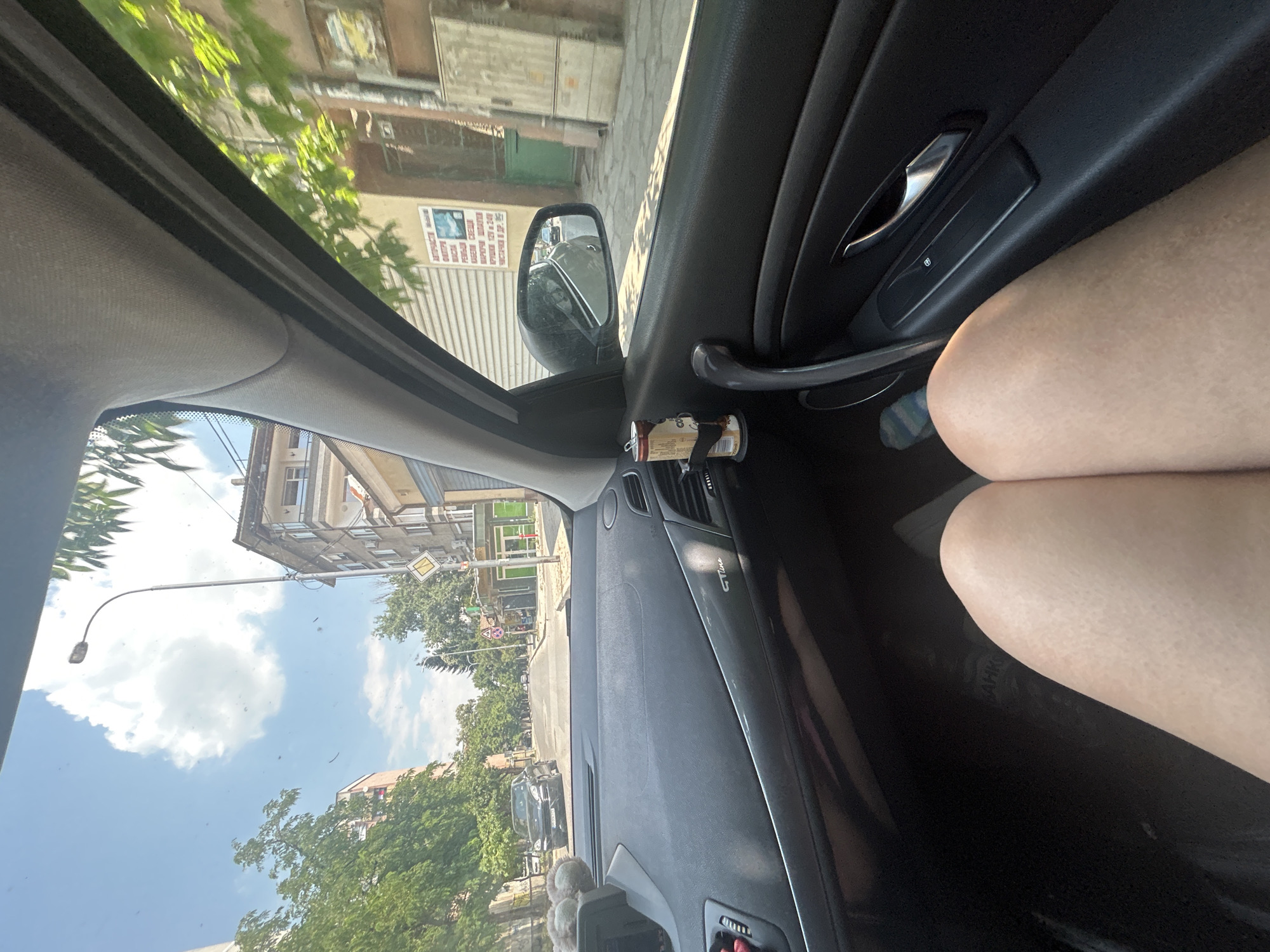 This media file depicts a view from inside a car, looking out the passenger side window and also showing the interior dashboard and a person's leg. The scene appears to be a street in a city, with buildings, trees, and a road visible. There are several cars on the road, and a traffic light is visible in the distance. The sky is blue with some clouds, suggesting it is daytime and the weather is fair.
Notable details include text on a sign outside the car which reads "АВТОЧАСТИ МАСЛА КАБЕЛИ СВЕЩИ МАРКУЧИ ШЛАЙФА КРУШКИ 12V 24V ЧИСТАЧКИ И ДР.", which translates to "Auto parts oils cables spark plugs hoses lubricants bulbs 12V 24V wipers and other". There is also text on the car's interior trim that reads "GT Line". A person's leg, presumably the passenger's, is visible in the foreground. The image is oriented vertically, as if taken by a person holding their phone that way. The overall impression is a snapshot of everyday life while traveling by car.