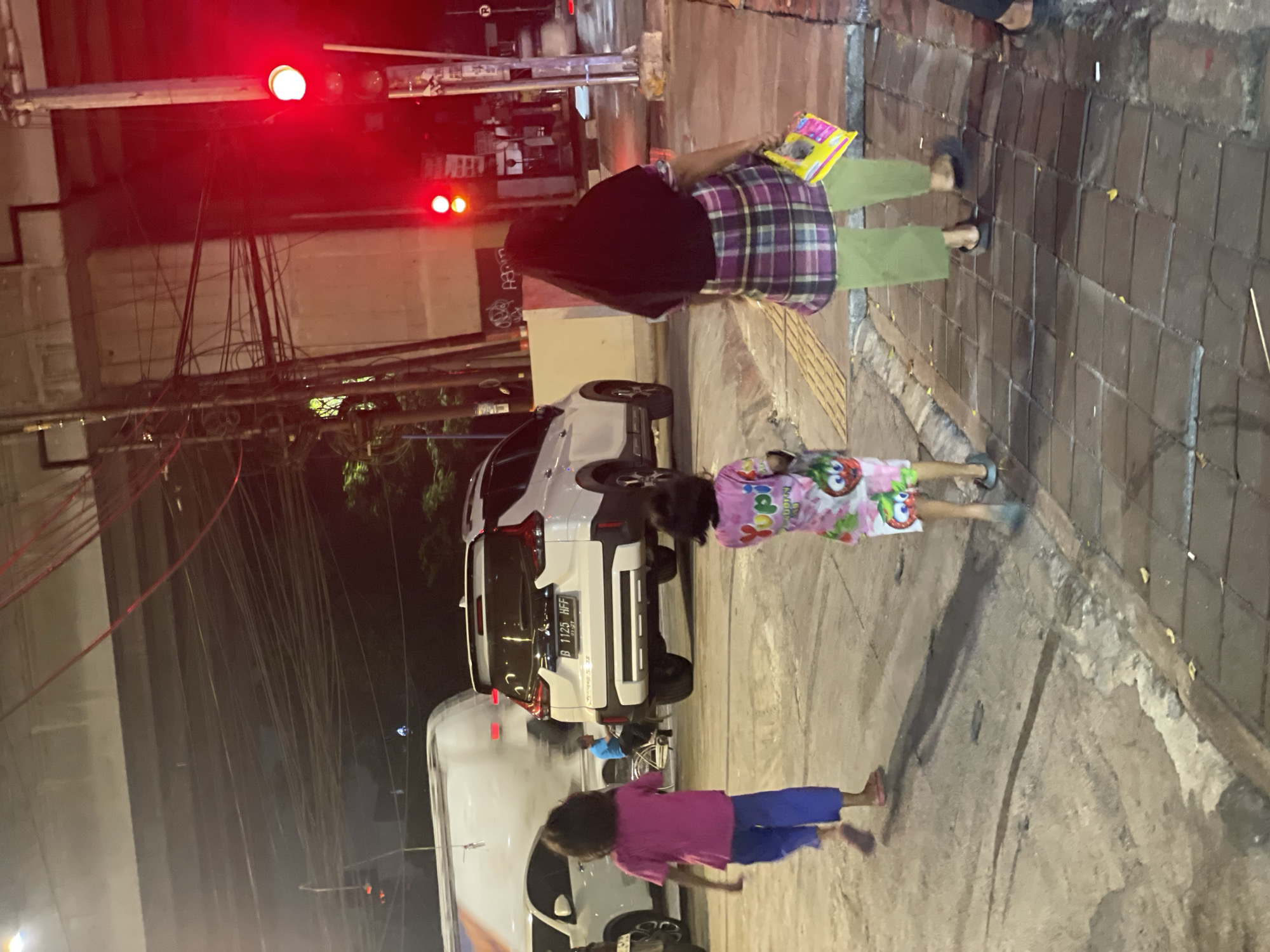 In North Jakarta, Indonesia, at dusk or night, a scene unfolds on a street illuminated by traffic lights and ambient lighting. The primary subjects are several people, including two children and an adult. The adult, wearing a dark headscarf and a plaid shirt over green pants, holds a colorful book or tablet. One child, dressed in pink pajamas with cartoon characters, stands near the adult. Another child, wearing a pink t-shirt and blue pants, is in the foreground.
The setting is a street with a sidewalk made of cobblestones and concrete. A white vehicle, possibly an SUV, is parked or stopped, its license plate visible as "B 1125 HFF." Another white vehicle, a larger van or truck, is also present. Overhead, a network of electrical wires crisscrosses the sky. The red traffic lights are a prominent feature, suggesting a controlled intersection.
The activity appears to be a moment of pause or waiting, perhaps for a traffic light to change or for movement. The children's stances suggest they are either standing still or in motion, possibly playing or waiting to proceed. The presence of the book or tablet with the adult implies an educational or recreational element.
Notable details include the time of day, indicated by the darkness and artificial lighting. The weather appears to be clear, with no signs of rain. The location cues, such as the vehicles and the general streetscape, are consistent with an urban environment in Indonesia. The emotions are not explicitly depicted but the scene appears neutral or calm. There is visible text on the license plate of the white vehicle.