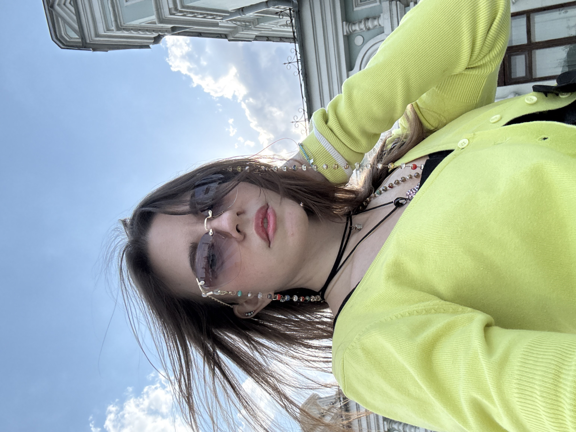 A young woman is seen from a low angle, likely a selfie. She wears sunglasses, a lime green cardigan, and multiple necklaces. Part of a building is visible in the upper left corner, as well as a blue sky with clouds. The building's architectural style suggests it is located in Moscow, Russia.