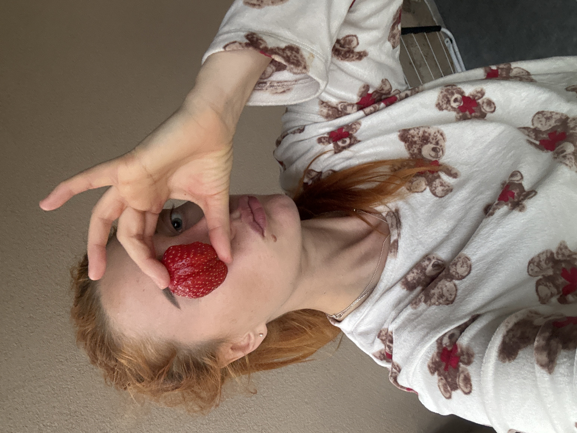 A woman with red hair and fair skin is holding a strawberry up to her face, between her eye and her mouth. She is wearing a white pajama top with a pattern of brown teddy bears with red bows. A silver necklace is visible around her neck. The background is a plain beige wall. There is no indication of the city of Budva, Montenegro in the image.