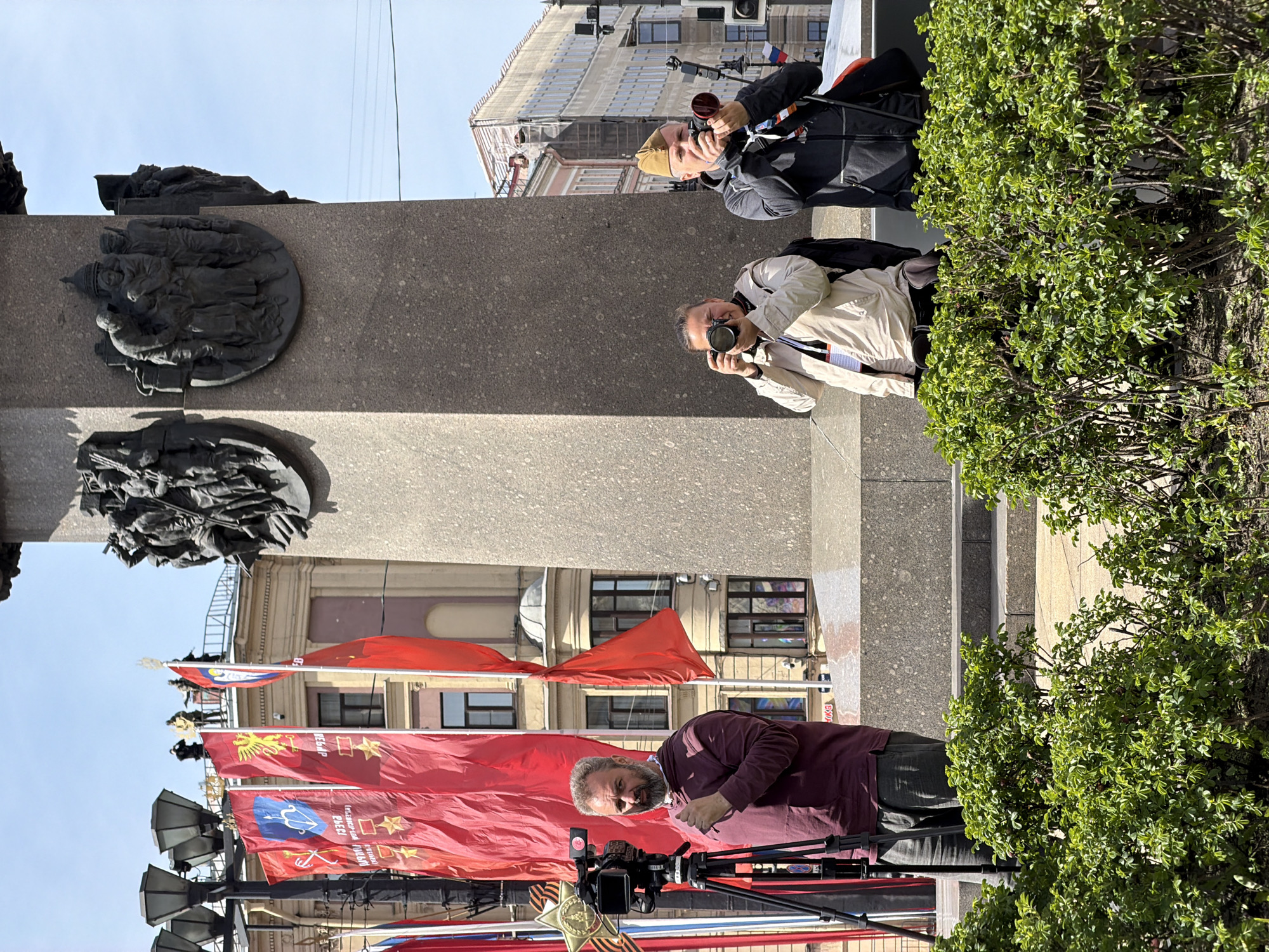 The image depicts a scene near the Victory Monument in Saint Petersburg, Russia, with a cameraman recording the area. Several red flags with Soviet symbols adorn the scene, along with decorative lighting. Two individuals are taking photos of the monument, a grey stone structure adorned with bas-relief sculptures. Lush green bushes flank the edge of the frame, while the sky is clear and blue. Buildings are visible in the background, suggesting an urban setting.