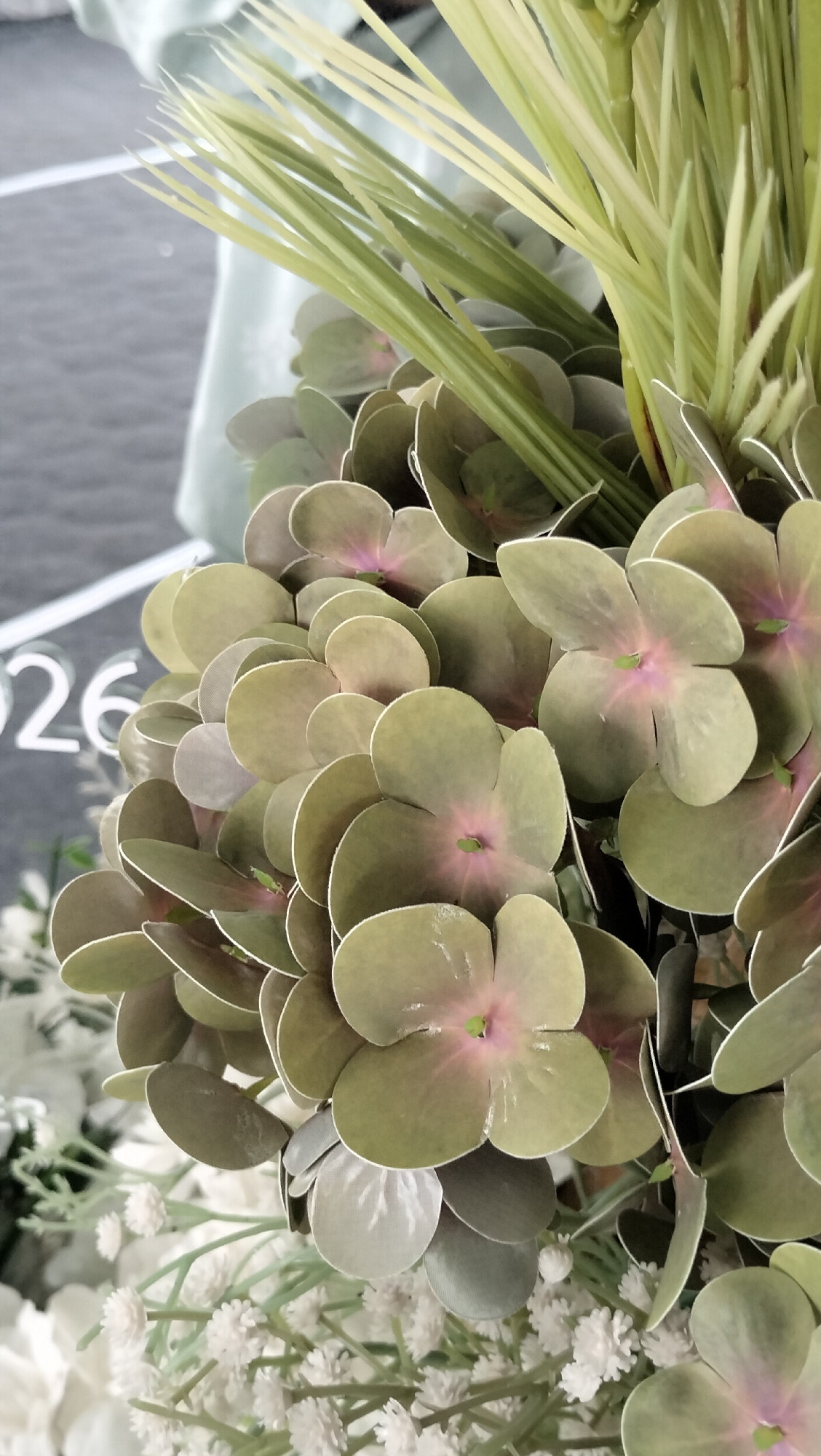 The image is a close-up, eye-level shot of artificial flowers. The main focus is on a cluster of muted green hydrangea-like flowers with hints of pink in their centers and lighter green, blade-like foliage behind them. To the lower left, smaller, delicate white flowers are visible, creating a softer texture. The background is blurred, showing what appears to be a light blue fabric on the left and a greyish, textured surface with white lettering that reads "026" on the left side of the frame. The lighting is soft and diffused, suggesting an indoor setting. The image does not depict any people, actions, or interactions. It is a still life composition of decorative floral elements. This scene is not identifiable as being in Lhokseumawe, Indonesia.