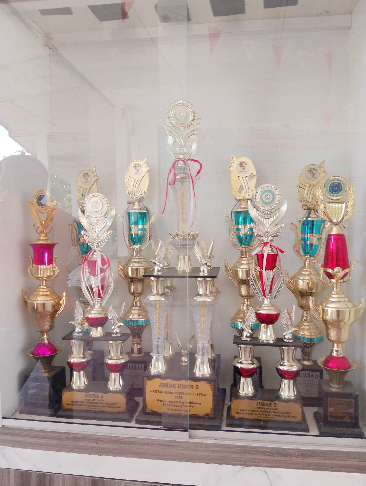 The image shows a display case filled with trophies of various sizes and designs, predominantly gold-colored with accents of red and teal. The trophies are arranged on black tiered stands. Several trophies have plaques attached to their bases, bearing inscriptions in Indonesian. One plaque reads "JUARA 3 CONCERT SHOW SIANTAR MARCHINGBAND FESTIVAL 2025 Memperingati Hari Pahlawan Se-Pematang Siantar 10 November 2020." Another reads "JUARA UMUM SIANTAR MARCHINGBAND FESTIVAL 2025". The display case appears to be in a public space, possibly an educational institution or community center, given the celebratory nature of the trophies. Reflections on the glass indicate an indoor environment with overhead lighting. The background is blurred but suggests a wall with some decorative elements. The location is identified as Serbelawan, Indonesia, implying the trophies are awards from a marching band festival held in or related to that area. No people are visible in the image.