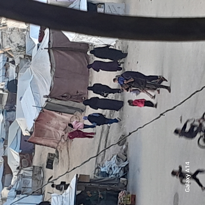 The image displays an overhead view of an outdoor scene, likely a street or alleyway within what appears to be a settlement or camp. The structures on both sides of the unpaved path are makeshift shelters, constructed from various materials like tarps, fabric, and possibly salvaged wood or metal, suggesting temporary or informal dwellings.
In the center of the path, several people are visible. A prominent feature of the scene is the presence of multiple individuals lying motionless on the ground. Starting from the left side of the path (when viewed upright), one person in a pink top and dark trousers is lying face down. Slightly to their right, another individual in a reddish-brown top and dark trousers is also lying face down. Further right, a group of people is gathered. There are at least five figures dressed in long, dark garments (possibly abayas or similar traditional attire), standing or bent over. Among them, one individual wearing a blue top and dark trousers is clearly visible, appearing to be attending to or observing another person who is lying on the ground near them. This person on the ground is mostly obscured but seems to be wearing dark clothing. Another individual in a red top and dark trousers is also lying face down further to the right of this central cluster.
In the background on the right side of the path, blurry figures are discernible. One appears to be riding a bicycle, and another seems to be running or walking. The foreground is partially obscured by a thick, dark, horizontal element, possibly a wire or structural beam, and a thinner diagonal wire crosses the mid-ground.
The time of day appears to be daytime, with visible shadows and bright areas indicating sunlight. The overall impression is one of a populated, possibly distressed, environment.
The text "Galaxy A14" is visible in the bottom right corner of the original image, indicating the device used to capture the photograph.