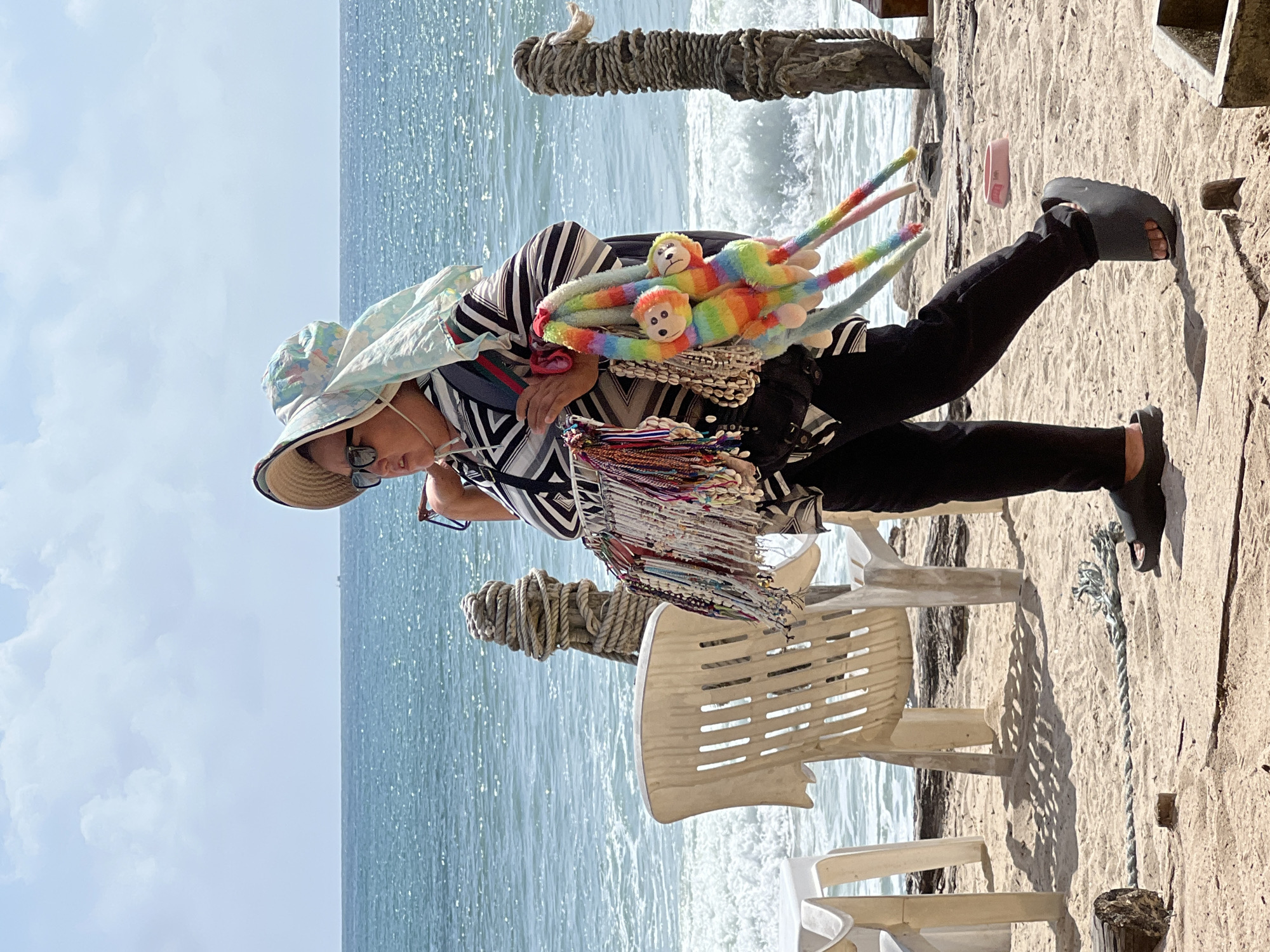 A person walks on a sandy beach in Choeng Thale, Thailand, near the ocean. The individual is wearing a wide-brimmed hat and sunglasses, a black and white striped shirt, and black pants. They are carrying a backpack and a selection of merchandise, including colorful necklaces and bracelets, as well as two rainbow-colored plush monkey toys. Several white plastic chairs and a wooden post wrapped with rope are visible on the beach, with the ocean waves breaking in the background under a partly cloudy sky.