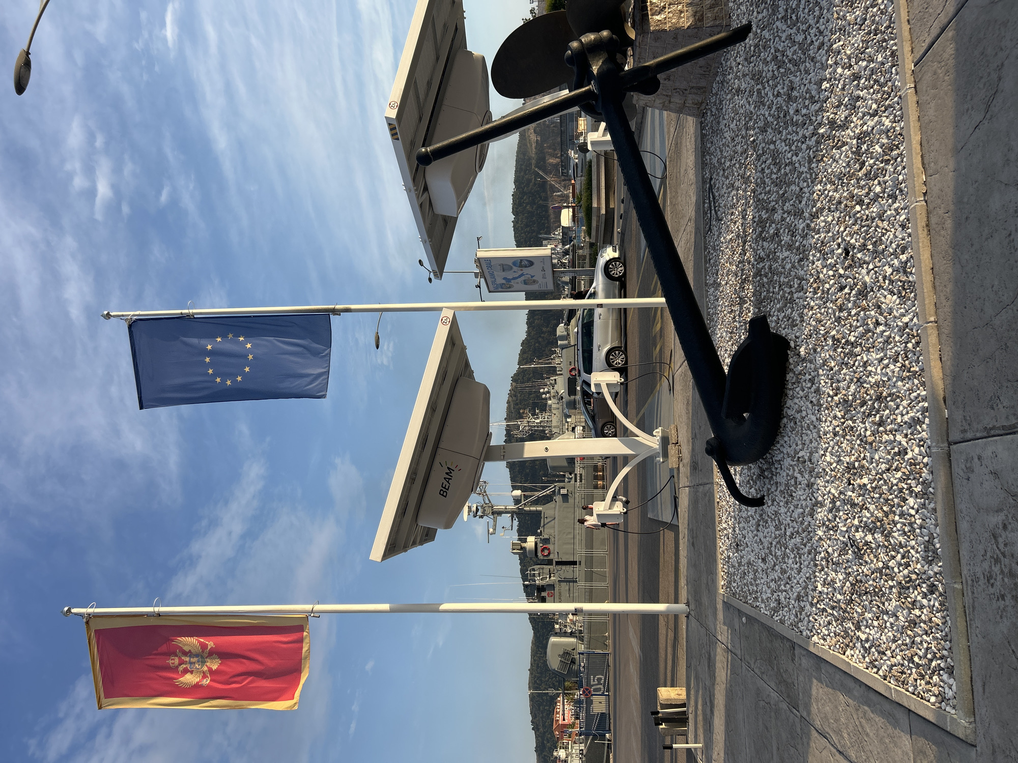 The image shows a harbor scene in Bar, Montenegro, under a clear blue sky with wispy clouds. Two flags are prominently displayed on flagpoles. The top flag is the blue European Union flag with its circle of stars. Below it is the red and gold flag of Montenegro, featuring a double-headed eagle crest.
In the foreground, a large, black anchor is mounted on a white pedestal, with its fluke resting on a bed of small white pebbles. The anchor appears to be decorative rather than functional, given its placement. Two white satellite dishes, labeled "BEAM," are mounted on white poles, facing upwards. These likely serve communication purposes for vessels in the harbor.
In the background, several boats and ships are docked. One white vehicle is visible on the paved area behind the anchor and satellite dishes. The scene is set in a harbor, suggesting a maritime environment. The lighting indicates it is daytime, likely late afternoon or early morning due to the warm light and long shadows. The overall impression is one of a well-maintained port facility, possibly with a tourist or commercial focus, given the presence of the EU flag. The clear sky suggests good weather.