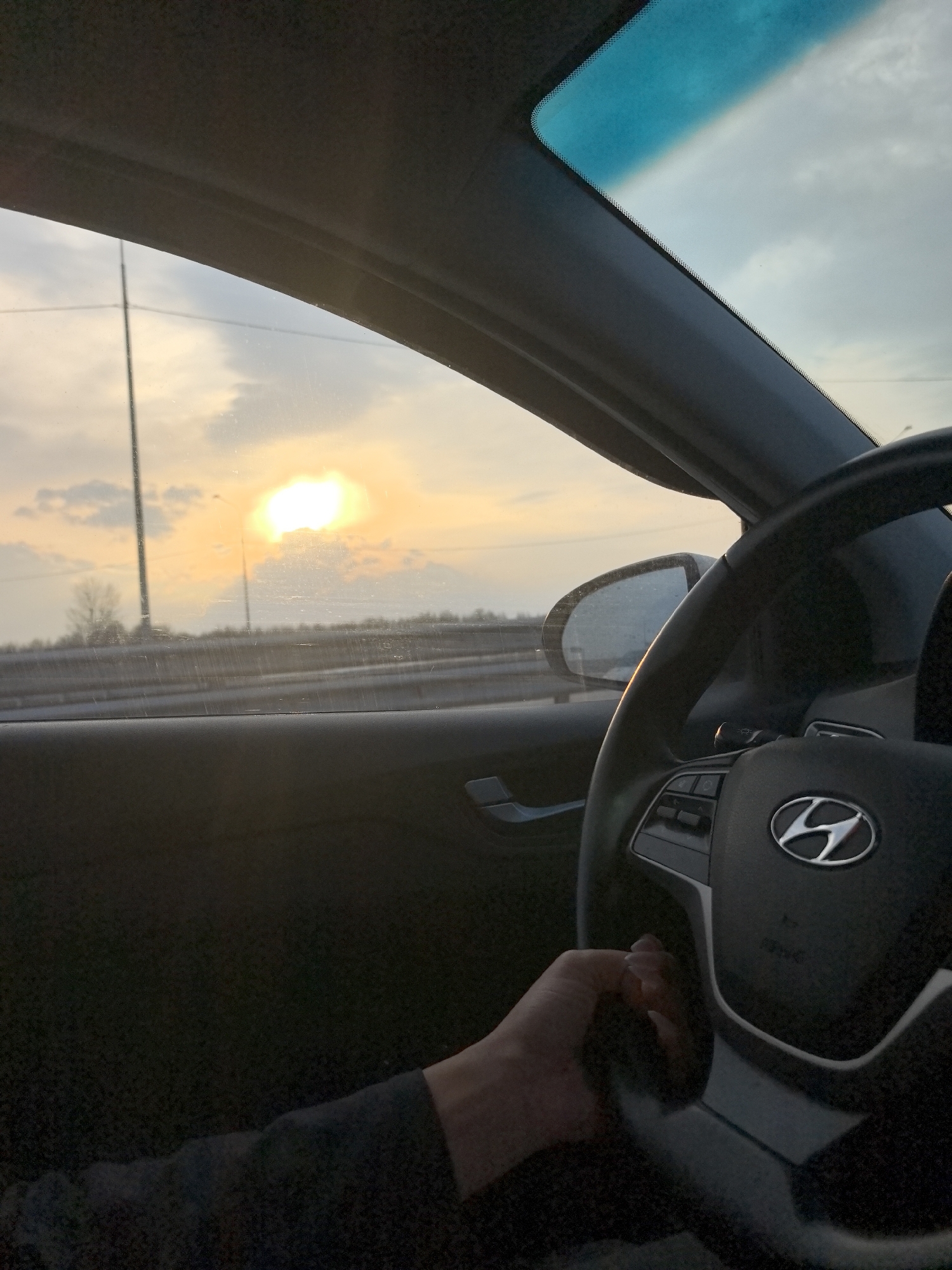 The image shows the interior of a car, specifically the driver's side. A hand is partially visible on the steering wheel, which features a Hyundai logo. The dashboard is black. Through the windshield, a view of the outside is seen: a highway or road with barriers, and in the distance, a bright sunset with clouds. A utility pole is also visible on the left side of the frame. The car appears to be in motion. The image does not provide sufficient information to determine the exact location in the city of Shali, Russia.