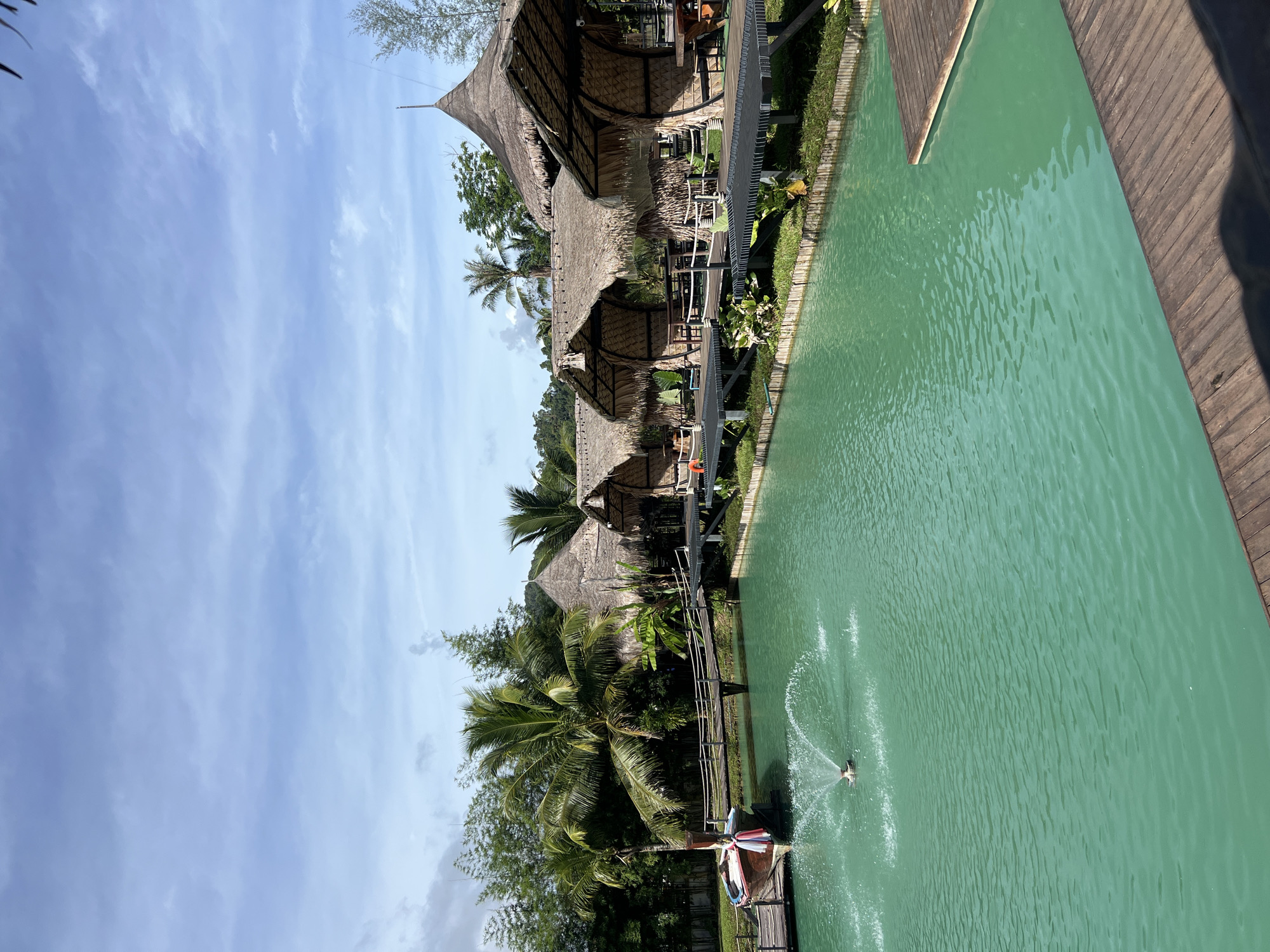 The image depicts a serene resort scene in Tambon Pa Kae Bo Hin, Thailand. The setting features traditional thatched-roof bungalows nestled amongst lush tropical foliage, including palm trees. A vibrant, turquoise swimming pool or lagoon is a prominent element, with a wooden deck bordering one side. The pool is enhanced by a fountain, creating a dynamic visual with water sprays.
In the foreground, a wooden jetty extends into the water, adorned with colorful decorative elements that resemble a pinwheel. A person is visible near the end of the jetty, possibly preparing to enter the water or enjoying the view. The sky above is a clear blue, punctuated by scattered white clouds, suggesting a pleasant, sunny day, likely in the late morning or early afternoon.
The overall impression is one of a tranquil and inviting tropical getaway. The architecture of the bungalows, with their natural materials and conical roofs, speaks to a local style, reinforcing the Thai location. The calm water and the presence of greenery contribute to a sense of relaxation and natural beauty. There is no visible text in the image.