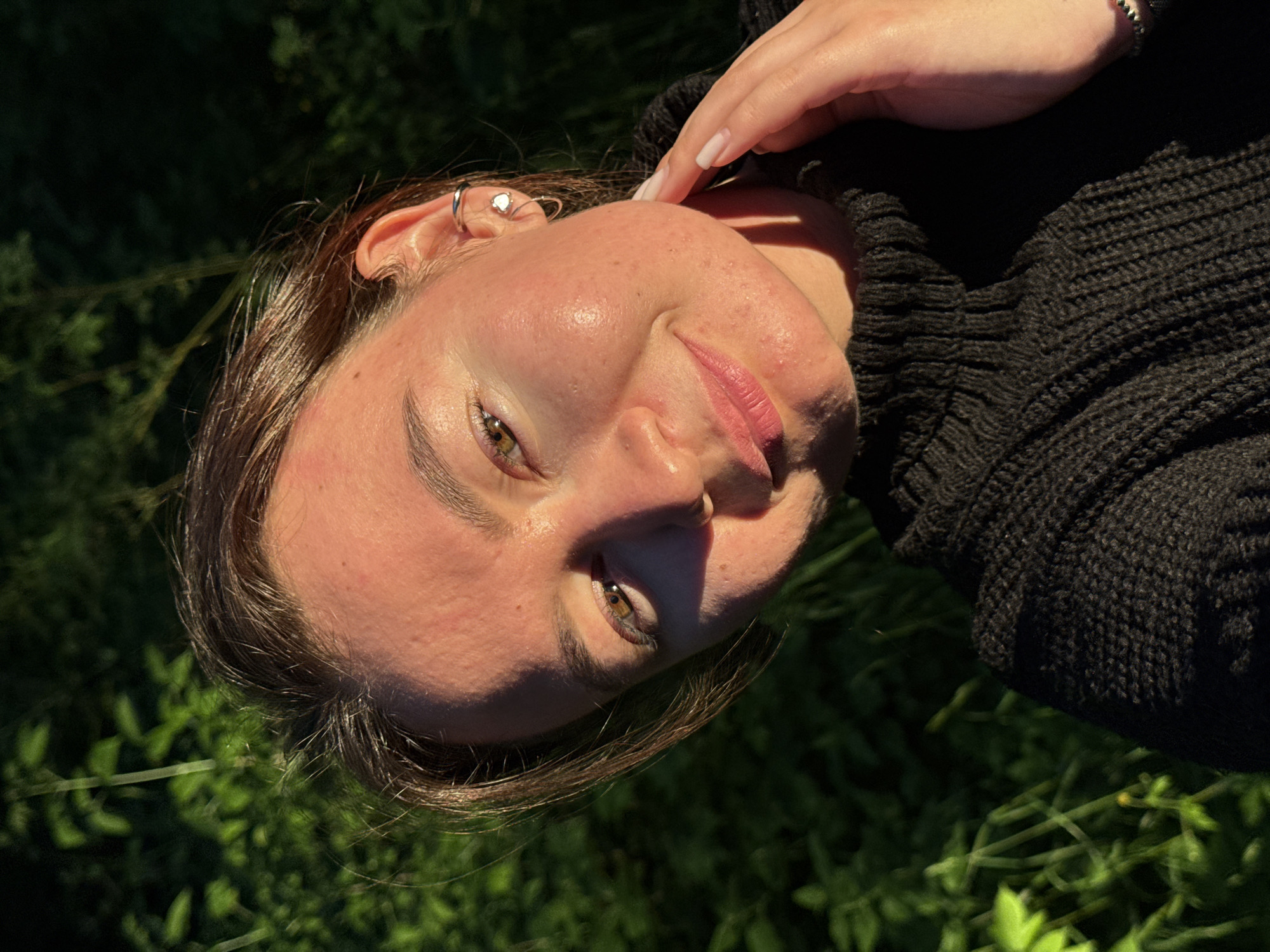 This is a close-up, outdoor portrait of a young person, looking directly at the camera with a gentle, soft smile. The head is tilted, suggesting a relaxed pose. The person has medium-brown hair and light-colored eyes, which appear to be hazel or brownish-green. Their fair skin shows natural texture, illuminated by direct sunlight.
The individual is wearing a dark, possibly black, ribbed or knitted sweater. On their left ear, a silver hoop earring and a small, heart-shaped stud earring are visible. A hand, with neatly manicured nails painted with light-colored polish, is gently placed near the temple/forehead area. A silver-toned beaded ring or bracelet is visible on the wrist of this hand.
The background consists of blurred, vibrant green foliage and grass, indicating an outdoor setting such as a park or garden, potentially in Chaikovsky, Russia. The lighting suggests late afternoon or early evening, often referred to as "golden hour," with warm, golden sunlight illuminating the left side of the face and hair, creating soft shadows on the right. The overall impression is one of calm and contentment. There is no visible text in the image.