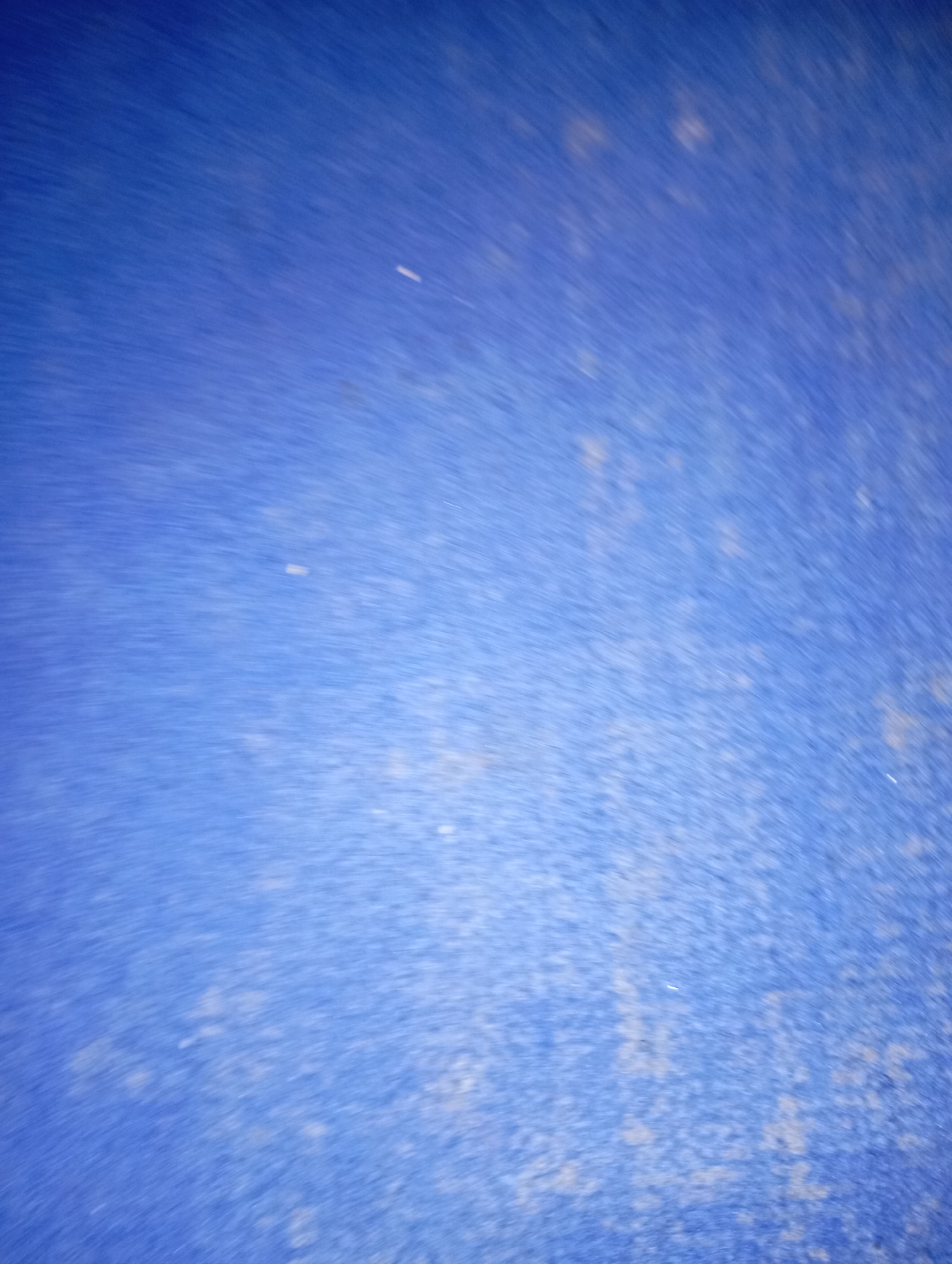The image is a close-up, abstract shot of a textured blue surface. The texture appears granular and possibly aged, with lighter, desaturated patches scattered across the predominantly vibrant blue background. There are subtle streaks and smudges, suggesting movement or a weathered finish. No specific location within New Stadium, Nigeria can be identified from this image. The scene is devoid of any discernible objects, people, or actions.