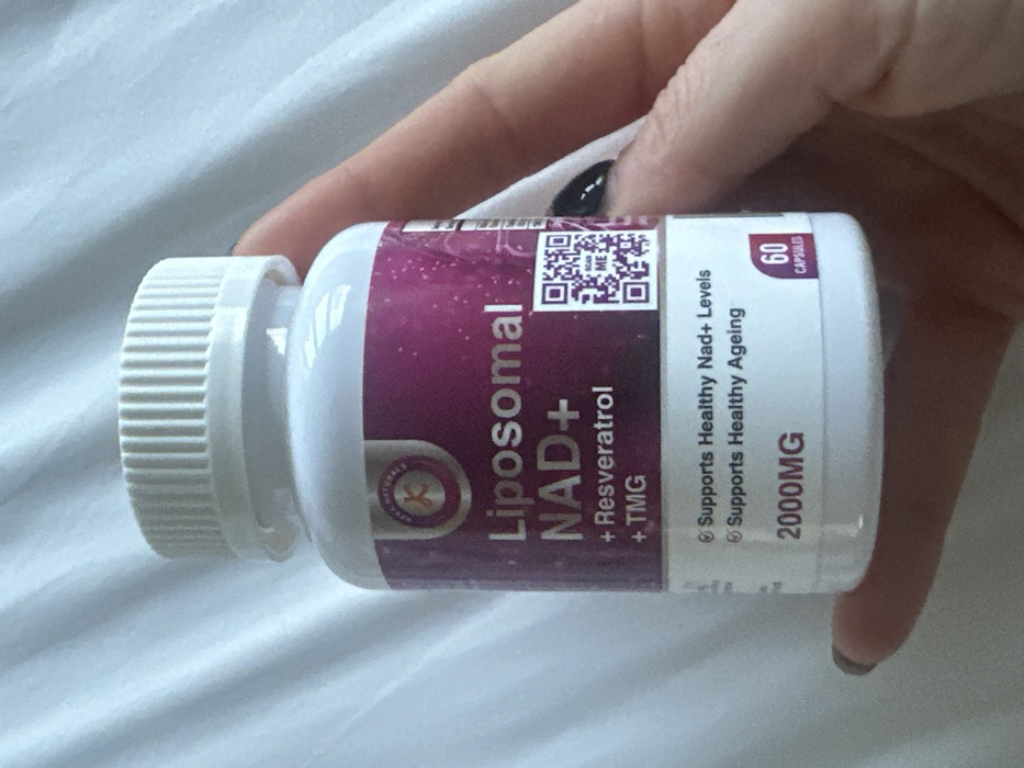 A human hand holds a white plastic bottle of "Liposomal NAD+ + Resveratrol + TMG" supplements. The bottle features a purple and white label from "KAYA NATURALS," indicating it "Supports Healthy Nad+ Levels" and "Supports Healthy Ageing," with a dosage of "2000MG" and containing "60 CAPSULES." A QR code with "SCAN ME" is present on the label. The hand, likely female, displays nails painted with dark polish, holding the bottle against a light-colored, textured fabric background, possibly a bedsheet or duvet. The specific city location within Wealden, United Kingdom, is not discernible from the image.