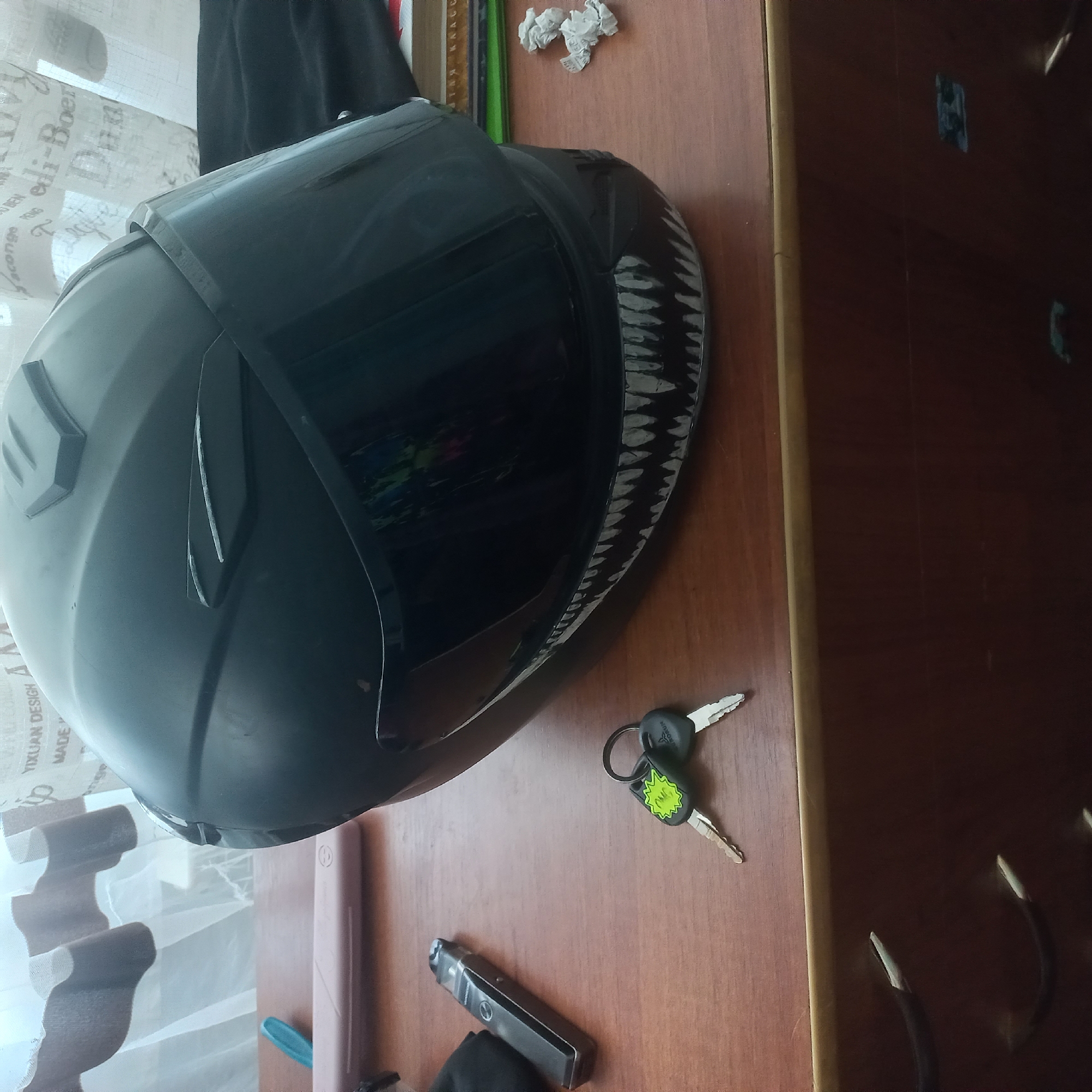 A black motorcycle helmet with a shark-tooth graphic on the visor rests on a wooden table. Beside the helmet are a vape pen and a set of keys with a bright green tag. In the background, a sheer curtain with a patterned design hangs in front of a window. The scene appears to be indoors, possibly in a bedroom or living area. There is no indication of the location in Selyshche, Ukraine, within the provided image.