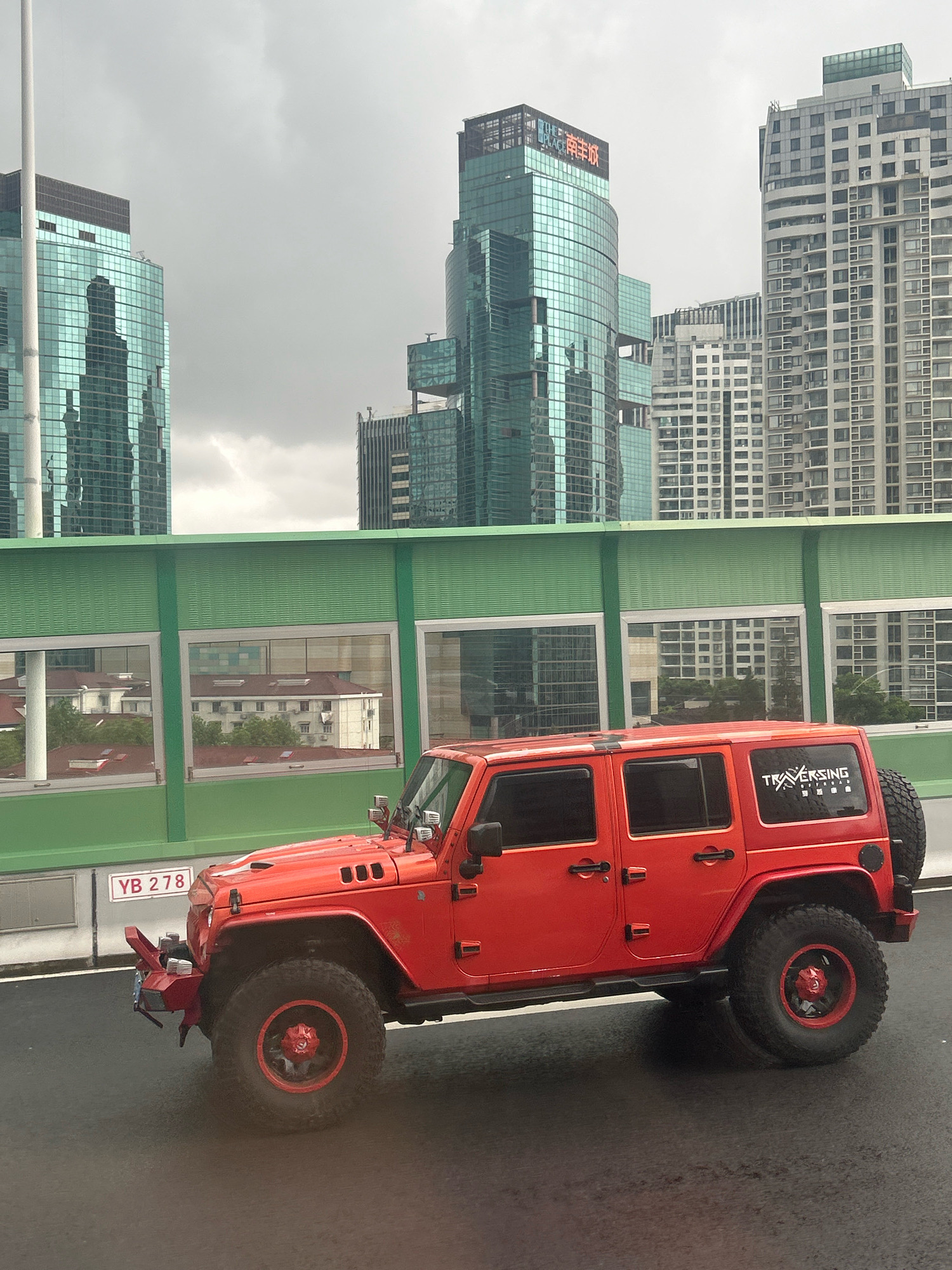 An orange Jeep Wrangler is parked on a wet road in front of a green, ribbed wall with windows. In the background, several tall, modern buildings with glass facades rise against a cloudy sky. One building prominently displays the characters "THE PLACE" and Chinese characters. A white license plate with the characters "YB 278" is visible on the front of the Jeep. The scene appears to be in an urban environment, possibly in the Changning District of China, given the architecture and signage. The lighting suggests an overcast day.