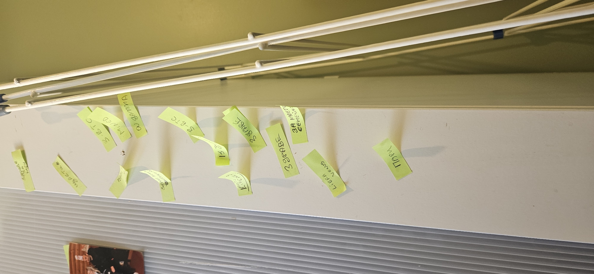 An upward-angled shot reveals a collection of yellow sticky notes affixed to a light-colored horizontal surface, possibly a shelf or a beam. Above this surface, several white metal rods of a drying rack are visible, casting shadows and partially obscuring some of the notes. Below the main surface, a textured gray panel, possibly a radiator or a blind, is seen, and a corner of a framed picture with people is visible on the far left. The wall above the shelf is a pale yellow-green.
The sticky notes are handwritten and arranged haphazardly, some curling at the edges, others overlapping. They display a mix of English and Cyrillic script.
Visible text includes:
- "158" (partially hidden)
- "Щастие" (Cyrillic for "Happiness")
- "4 MEH"
- "3-BTC" (appearing twice)
- "MINED"
- "ON GAMMA"
- "LIBRA VENUS"
- "3 GRADE" (appearing multiple times; some OCR identifies "S&PABE", "39PABE", or "3 драве," suggesting variations in handwriting or Cyrillic characters)
- "3A MOET"
- "CEMETG" (OCR suggests "CEMEGS")
- "Пари" (Cyrillic for "Money")
The mixture of terms like "BTC" (Bitcoin), "MINED," "LIBRA VENUS," and the Cyrillic words suggests a personal collection of notes with diverse themes, possibly relating to cryptocurrency, astrology, and general life concepts. The image is an indoor shot, with no discernible time of day or weather. According to the provided context, the location is Norwich, United Kingdom.